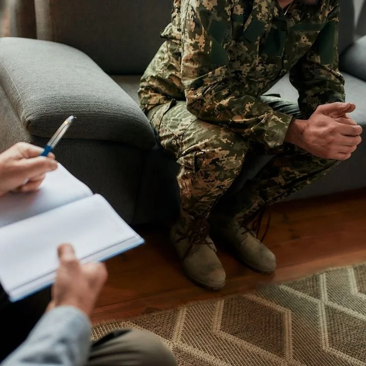 A person in military camouflage uniform is sitting on a couch with hands clasped, during a discussion or interview with someone taking notes.