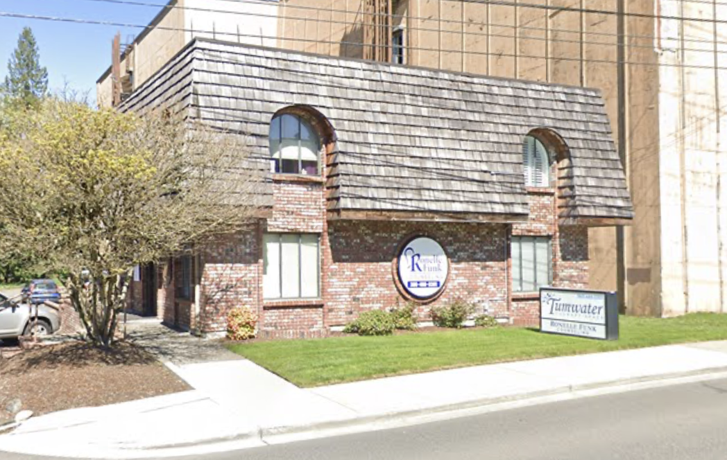A small brick commercial building with a shingled roof and arched windows. There are signs on the lawn, one reads 'Roseville Funk' and the other 'Tinkerwater Bottled Water.' A parking lot with cars is visible nearby, and a large building or wall is in the background.
