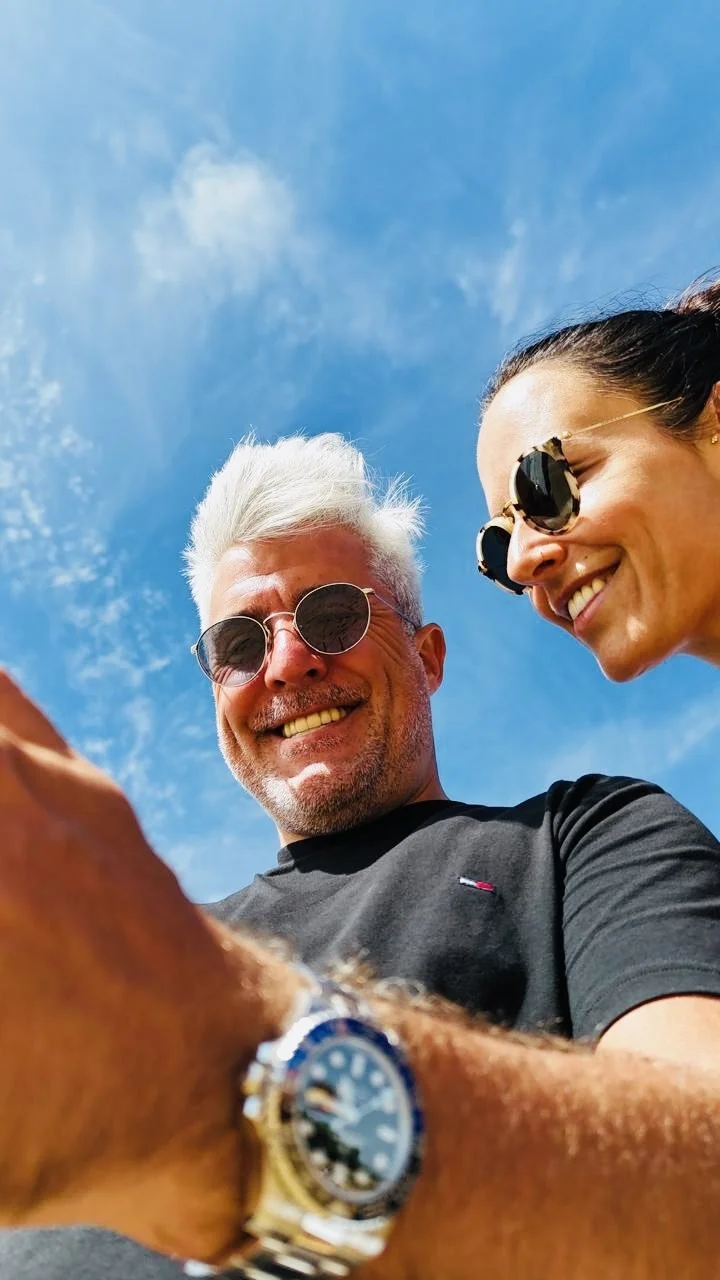 Two smiling people, a man with gray hair and sunglasses and a woman with dark hair and sunglasses, looking down at a camera or phone, with a blue sky with some clouds in the background.