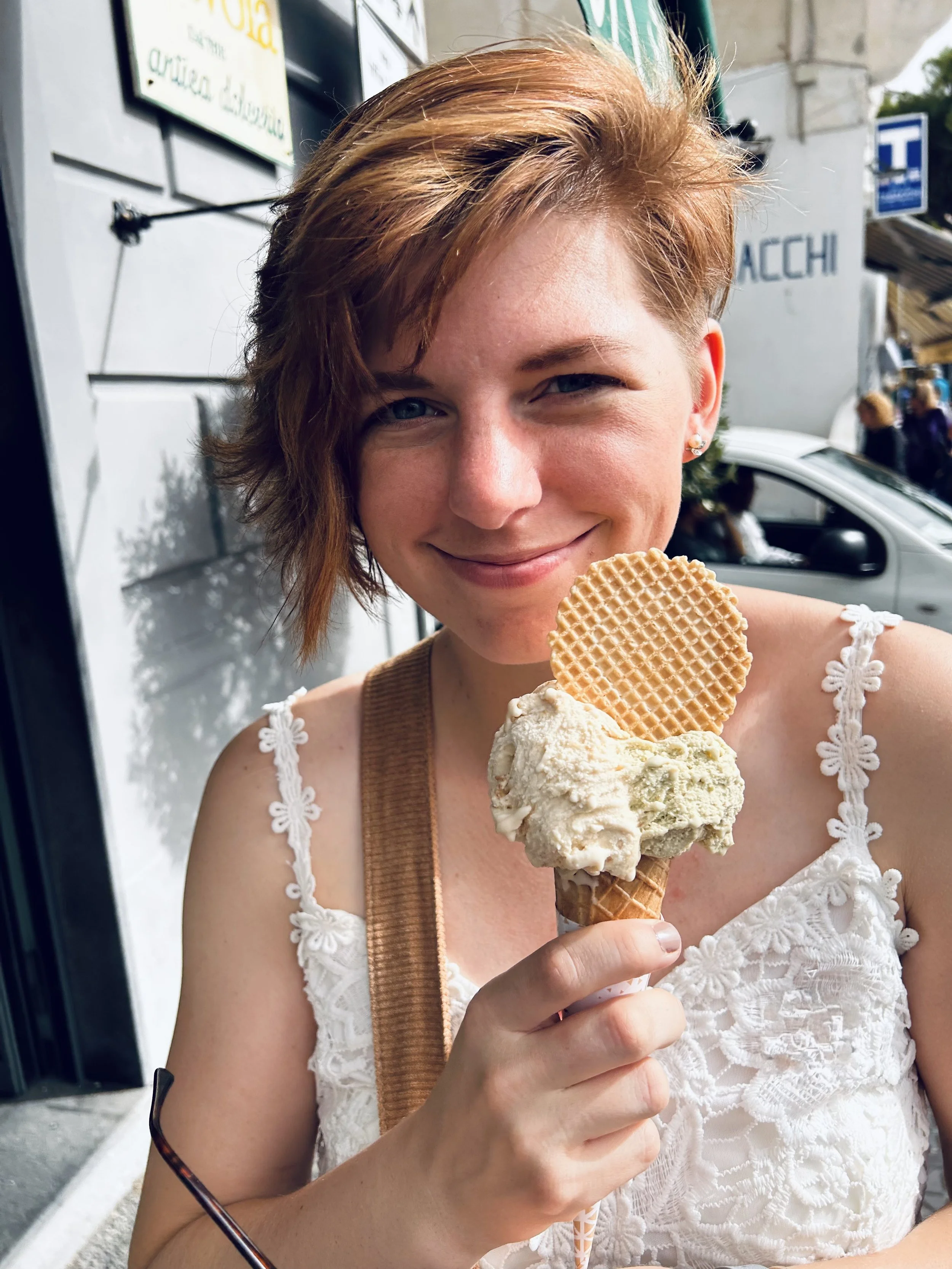 A woman with short, reddish hair and a white lace top is holding a large ice cream cone with two scoops, one vanilla and one pistachio, topped with a round cookie.