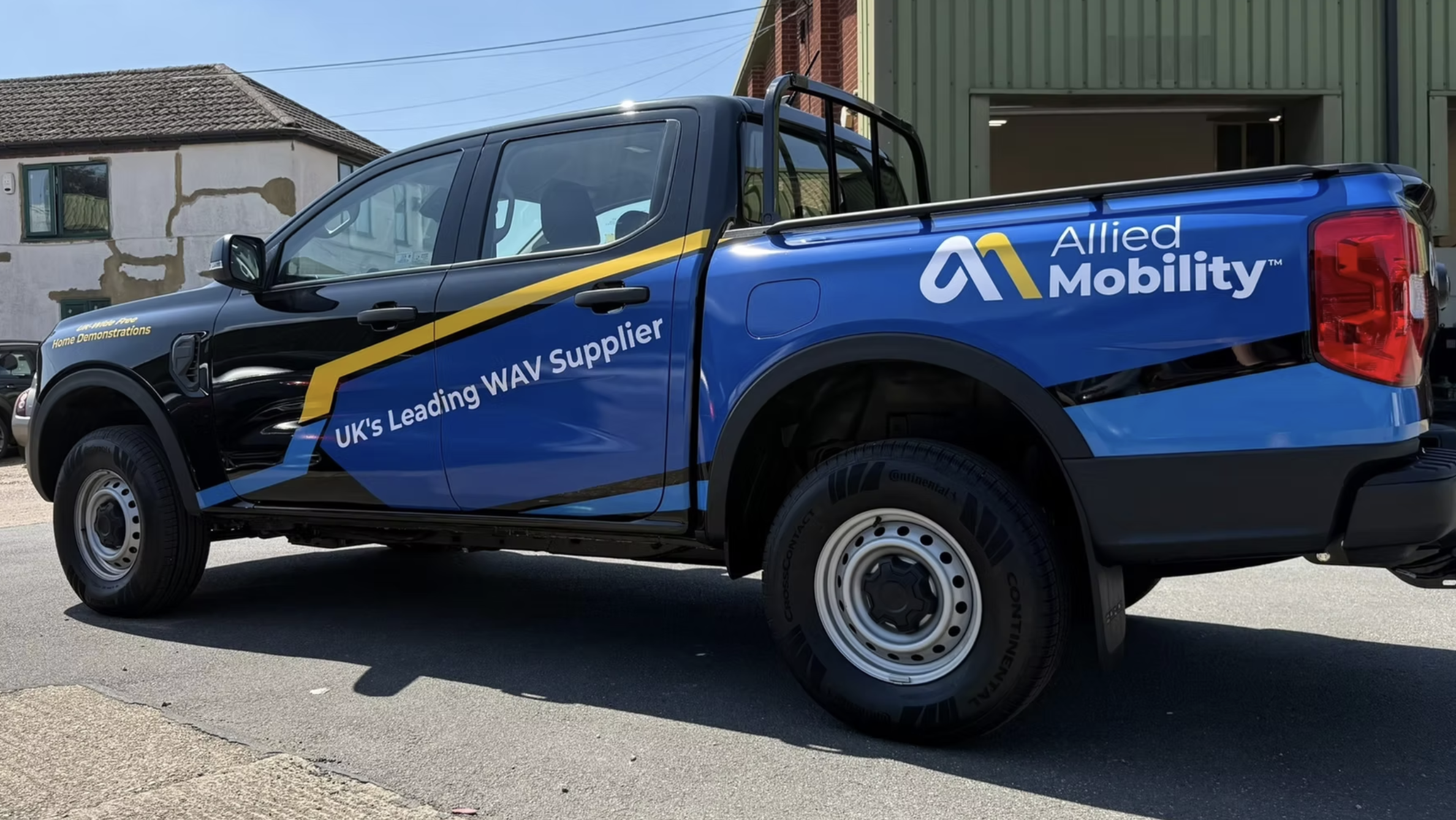 A blue pickup truck with branding for Allied Mobility and WAV Supplier parked on a street. The truck has a yellow diagonal stripe and text indicating it is a UK-based mobility service.