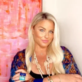A blonde woman with blue eyes, wearing a colorful patterned blazer, multiple pearl necklaces, and jewelry posing in front of a pink abstract painting.
