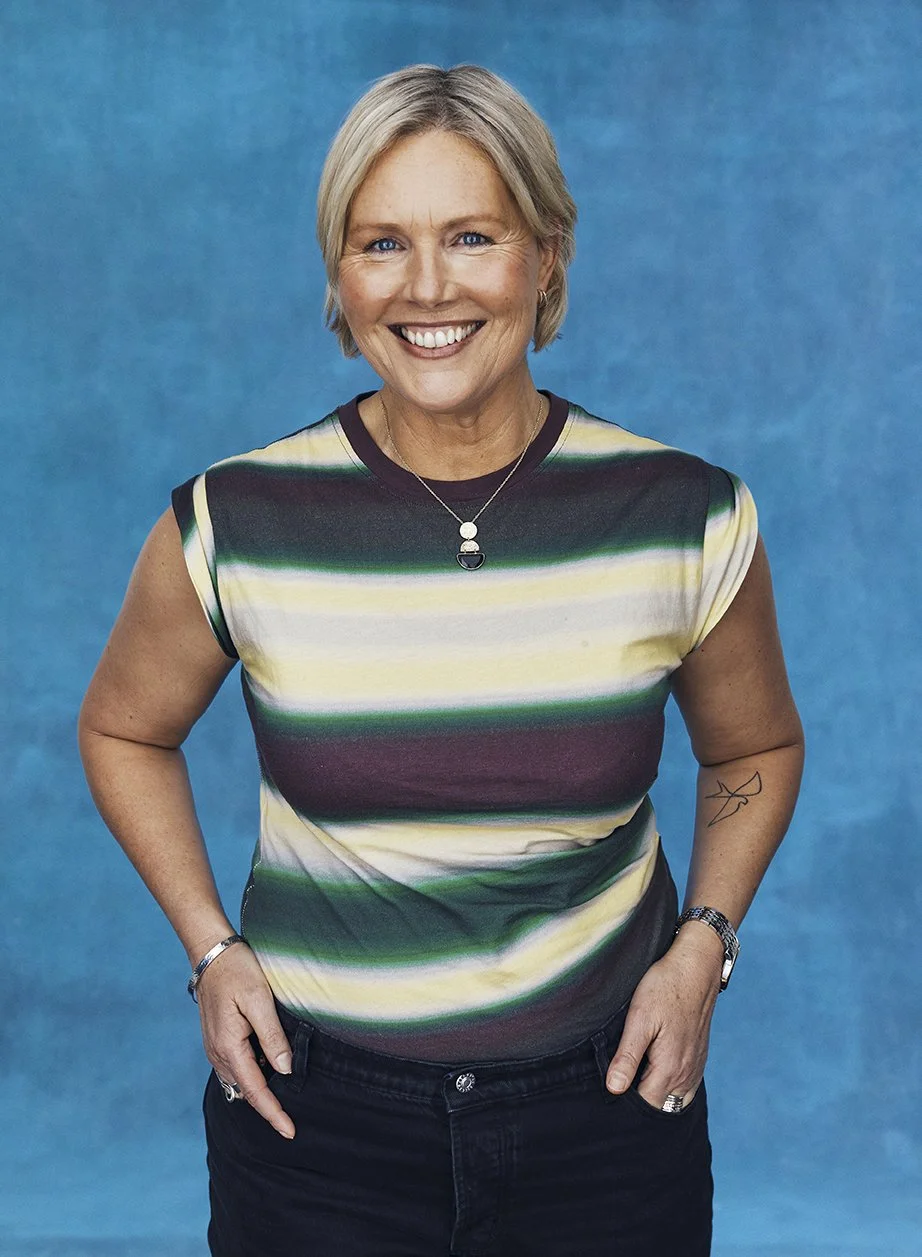 A smiling woman with short blonde hair, wearing a multicolored striped sleeveless top and black pants, standing against a blue background.