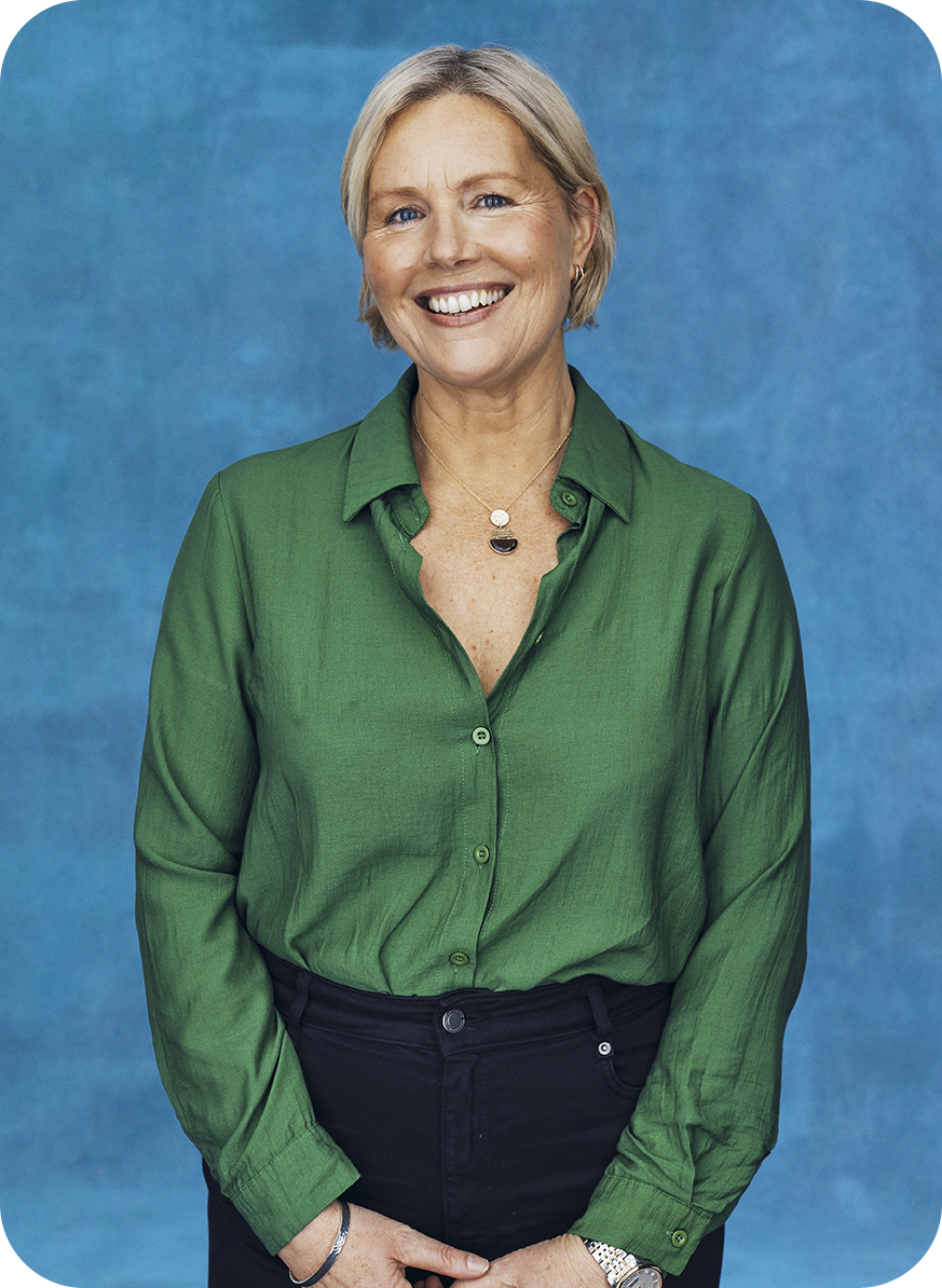 An older woman with short gray hair, smiling, wearing a green button-up shirt, black pants, a watch, a bracelet, and a necklace, standing against a blue background.