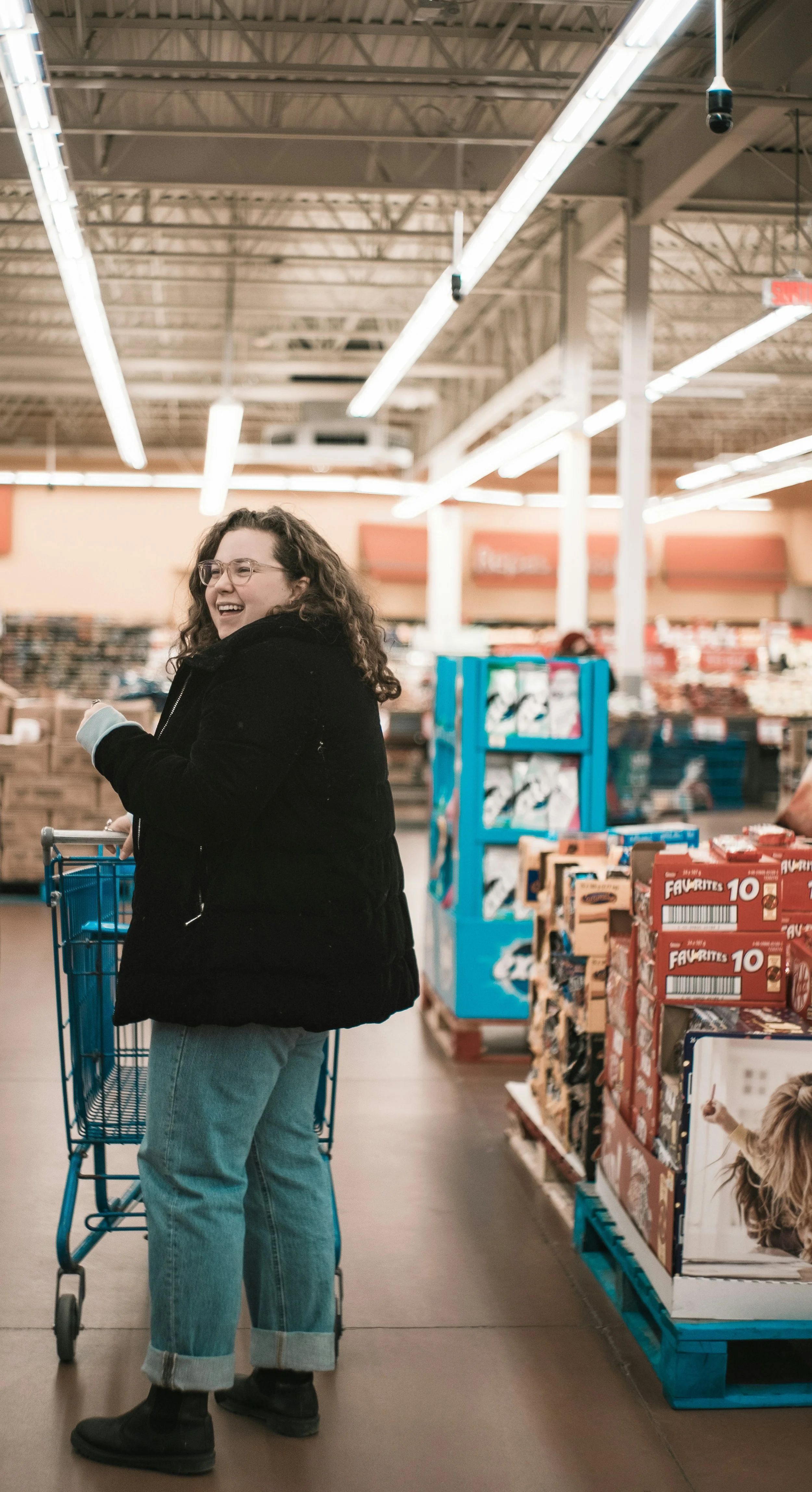 A woman with curly hair wearing glasses, a black jacket, and light blue jeans with rolled cuffs, smiling while shopping in a grocery store, pushing a blue shopping cart.