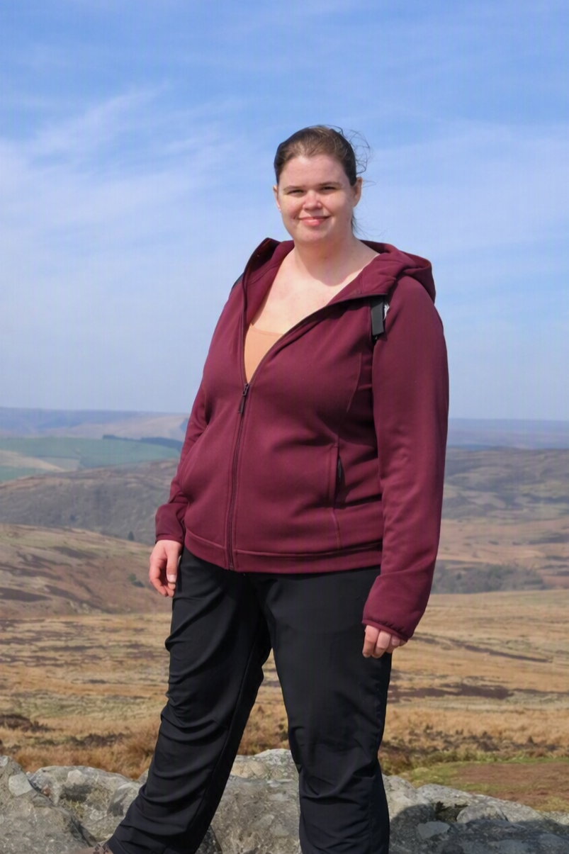 A woman standing on a hilltop with a vast landscape in the background, wearing a maroon zip-up jacket and black pants.