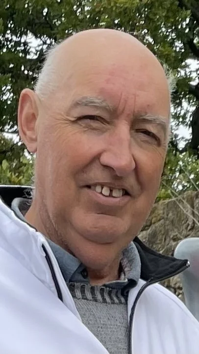 A close-up of a smiling older man with a bald head, wearing a light-colored jacket and a plaid shirt, outdoors near trees.