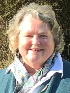 A smiling woman outdoors with gray curly hair, wearing a blue jacket and a patterned scarf.