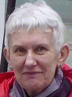 A person with short white hair and light skin smiling at the camera, wearing a purple jacket and red scarf.