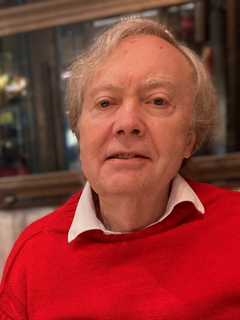 A man with light-colored hair and a slightly serious expression, wearing a red sweater over a white collared shirt, sitting indoors with a blurred background.