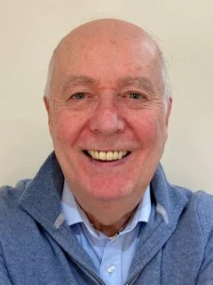 A smiling elderly man with a bald head, wearing a blue sweater over a collared shirt, standing against a plain background.