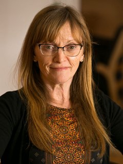 A woman with long, straight, light brown hair and glasses, wearing a dark top with a patterned shirt underneath, smiling at the camera.