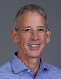 A middle-aged man with gray hair and glasses smiling, wearing a blue collared shirt against a gray background.