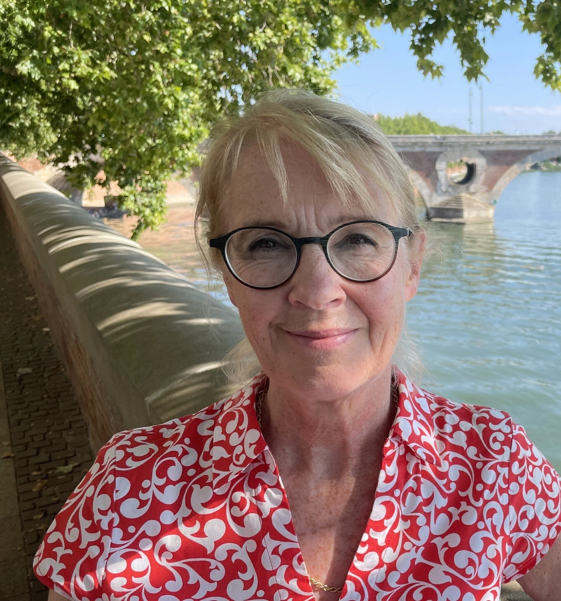 Close-up photo of a woman with short blonde hair and glasses, wearing a red and white patterned shirt, smiling outdoors near a body of water with a bridge in the background, shaded by green trees.