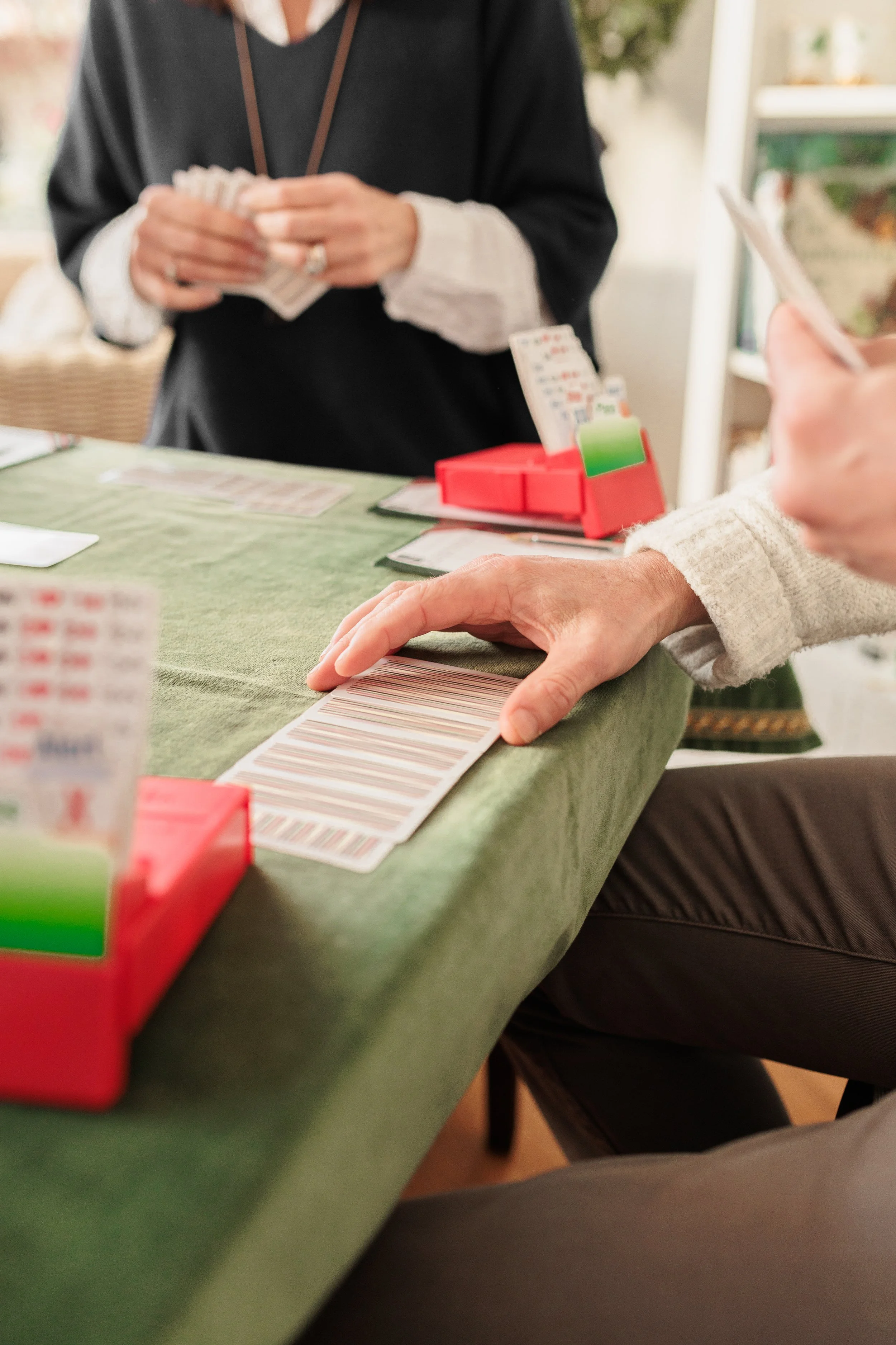 People playing bingo at a table with bingo cards, a red bingo chip tray, and a paper card. One person is holding a bingo card and the other is reaching for their own.
