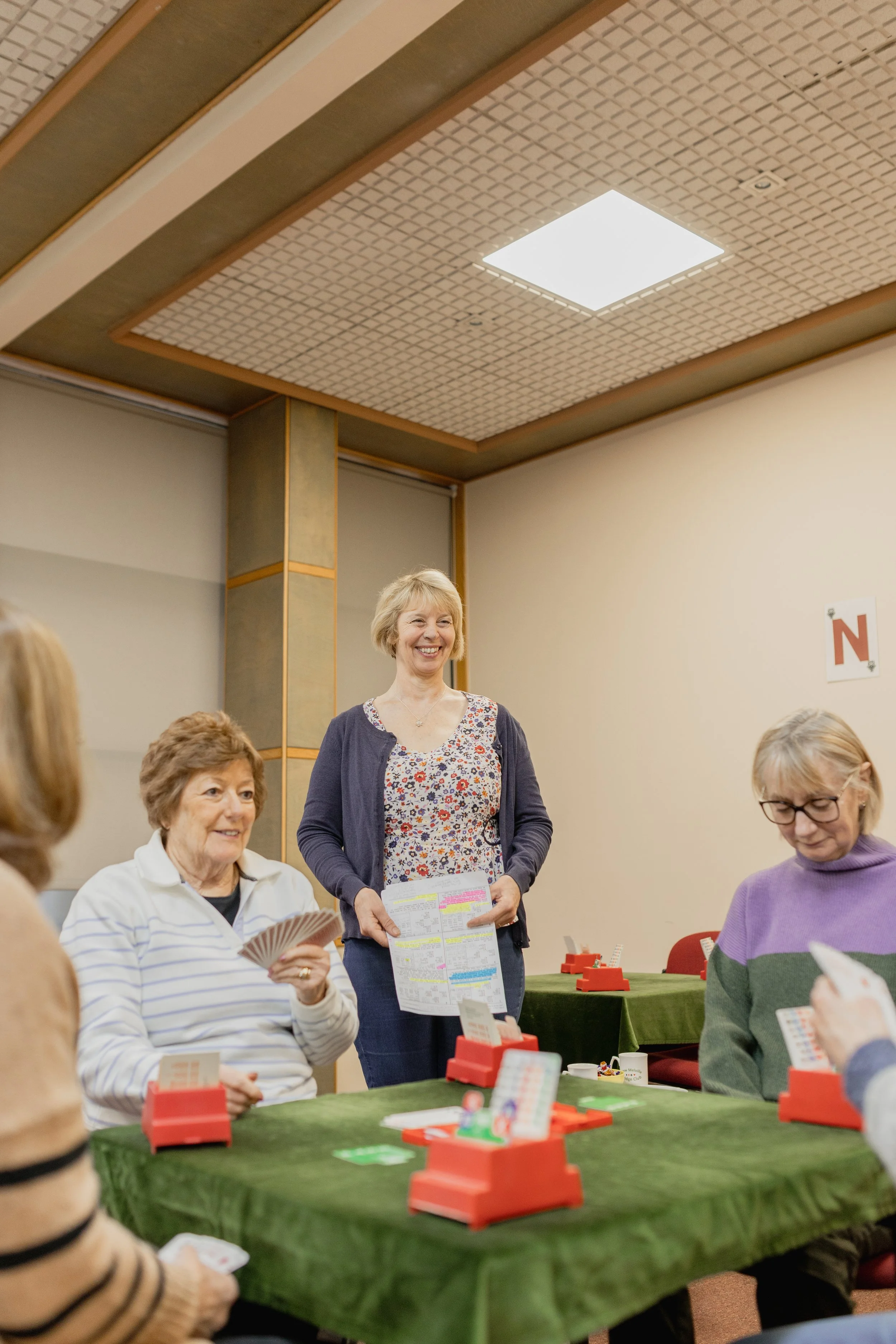 A group of women playing a board game at a table covered with a green cloth in a room with beige walls and a ceiling with a light fixture.