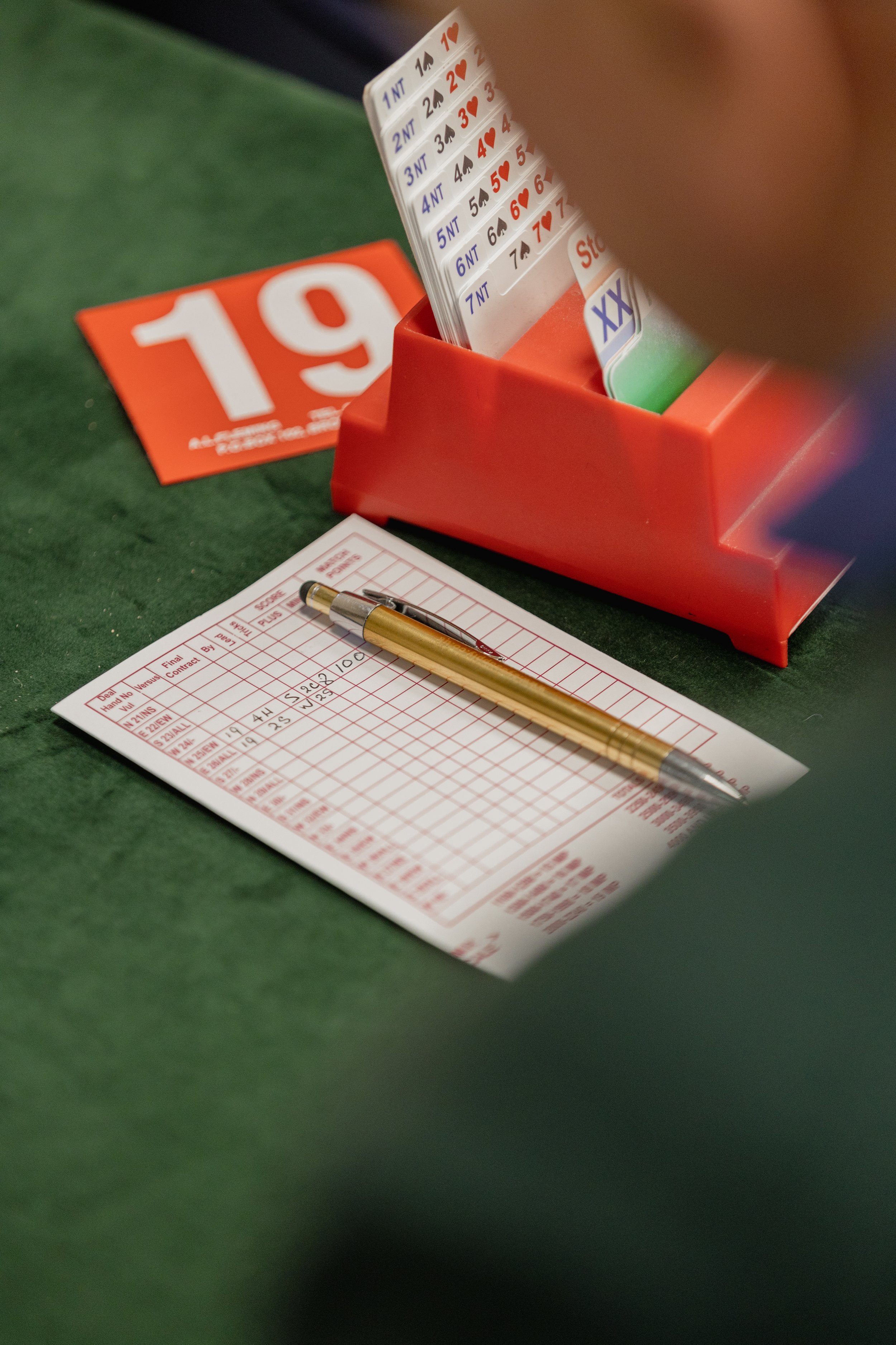 A betting slip with a gold pen on it, a red and white bingo card, a red number 19 sign, and a red ticket rack with poker cheat sheets in it, all on a green felt surface.