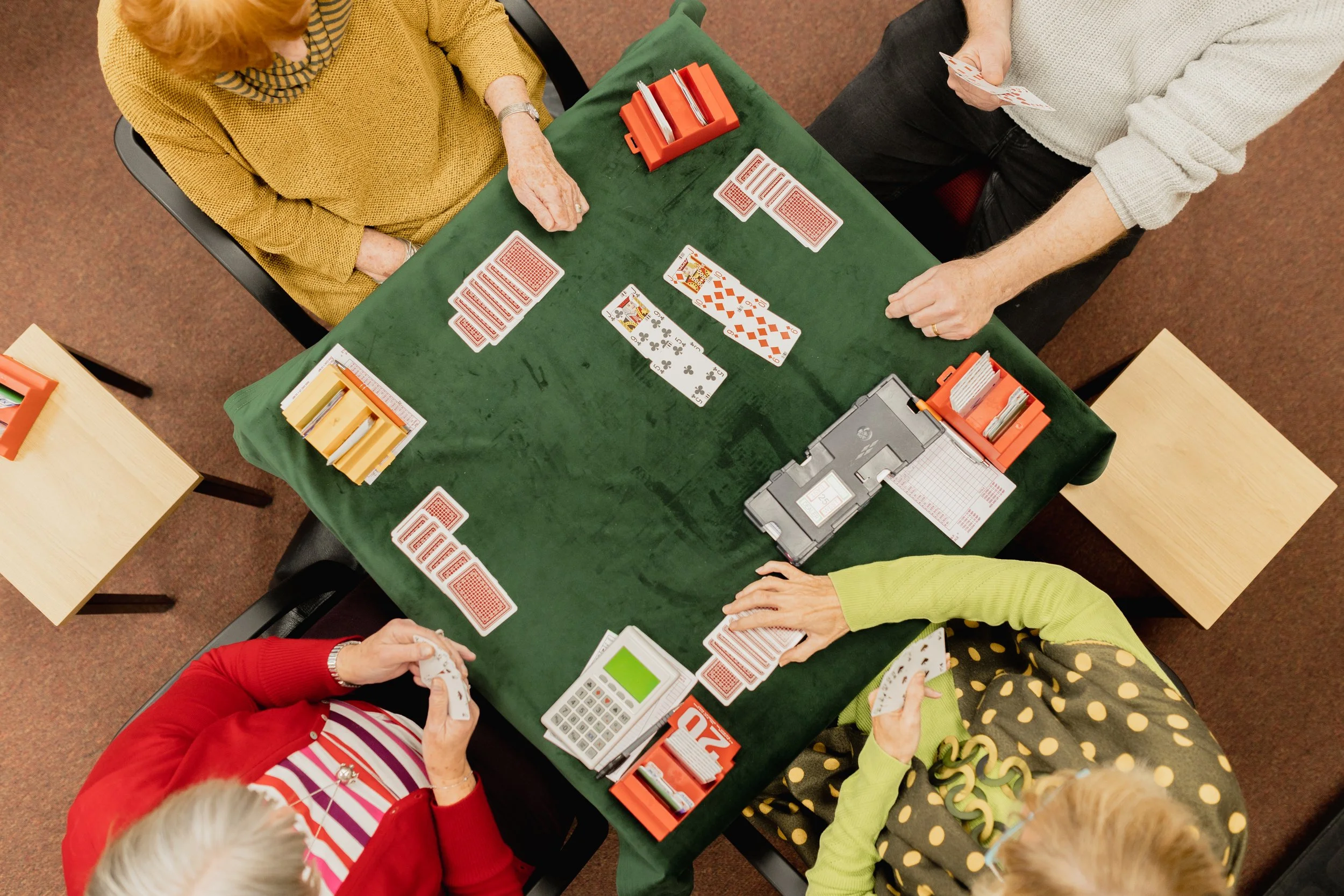 People playing a game of cards at a green-covered table, with some holding cards and others looking at the cards on the table, in a room with brown carpeted floor.