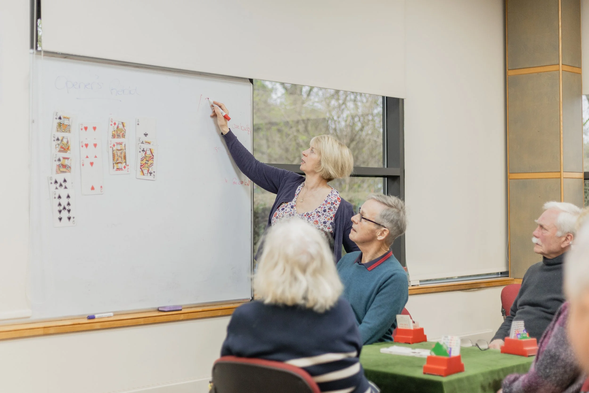 A group of older adults in a classroom, watching a woman standing at a whiteboard with playing cards attached, giving a presentation or lesson.