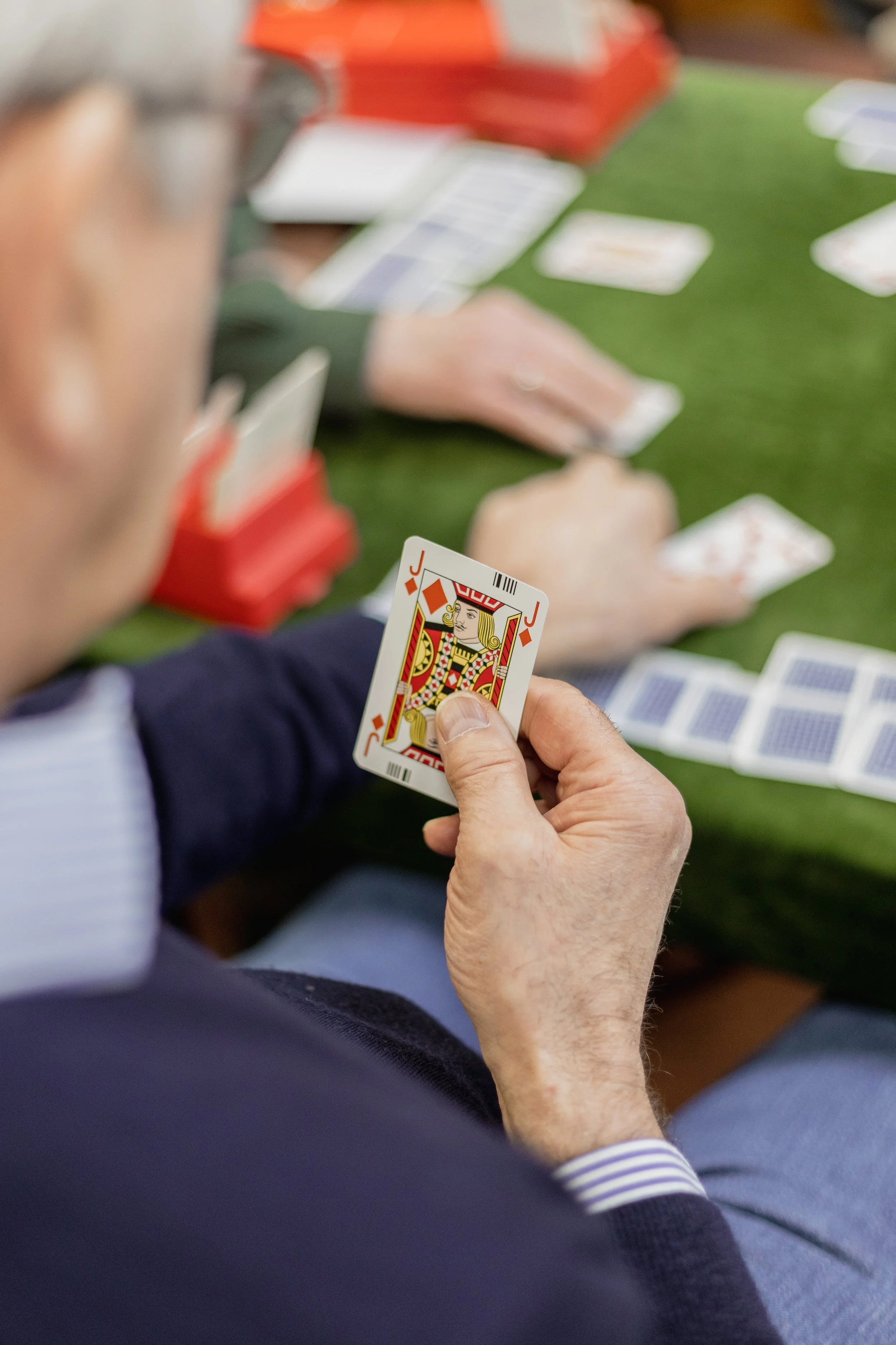 Close-up of a person holding a playing card, the Jack of Diamonds, during a card game. The person's hand is visible, and they are seated at a table with cards and game pieces. The person's arm is in focus, showing a dark sleeve with a striped shirt cuff. The background shows a blurry tabletop with other playing cards and game materials.