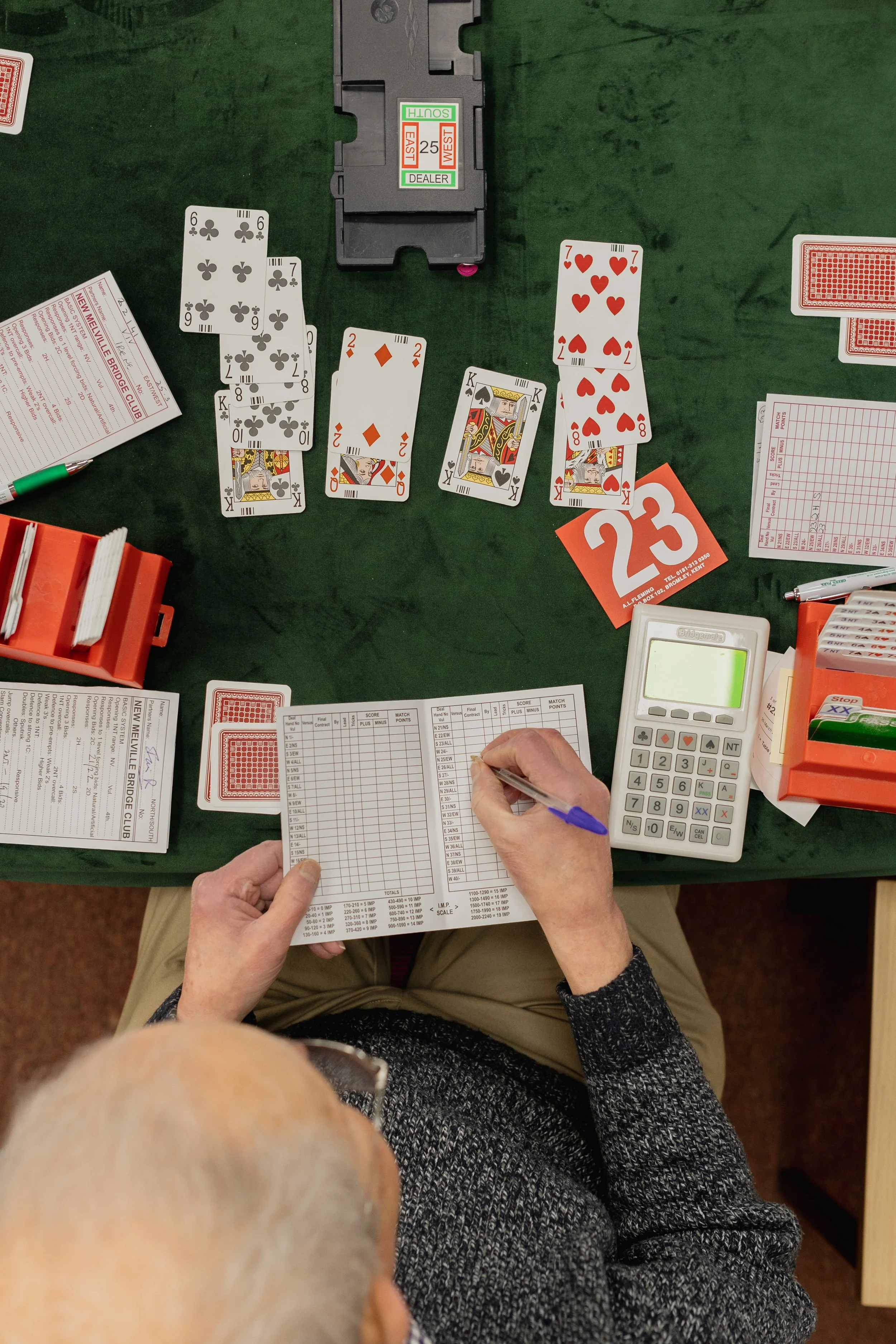 Two elderly men are playing a game of bridge at a green felt table. One man is writing on a score sheet while the other looks on. There are playing cards, score sheets, and a card dealer box on the table.