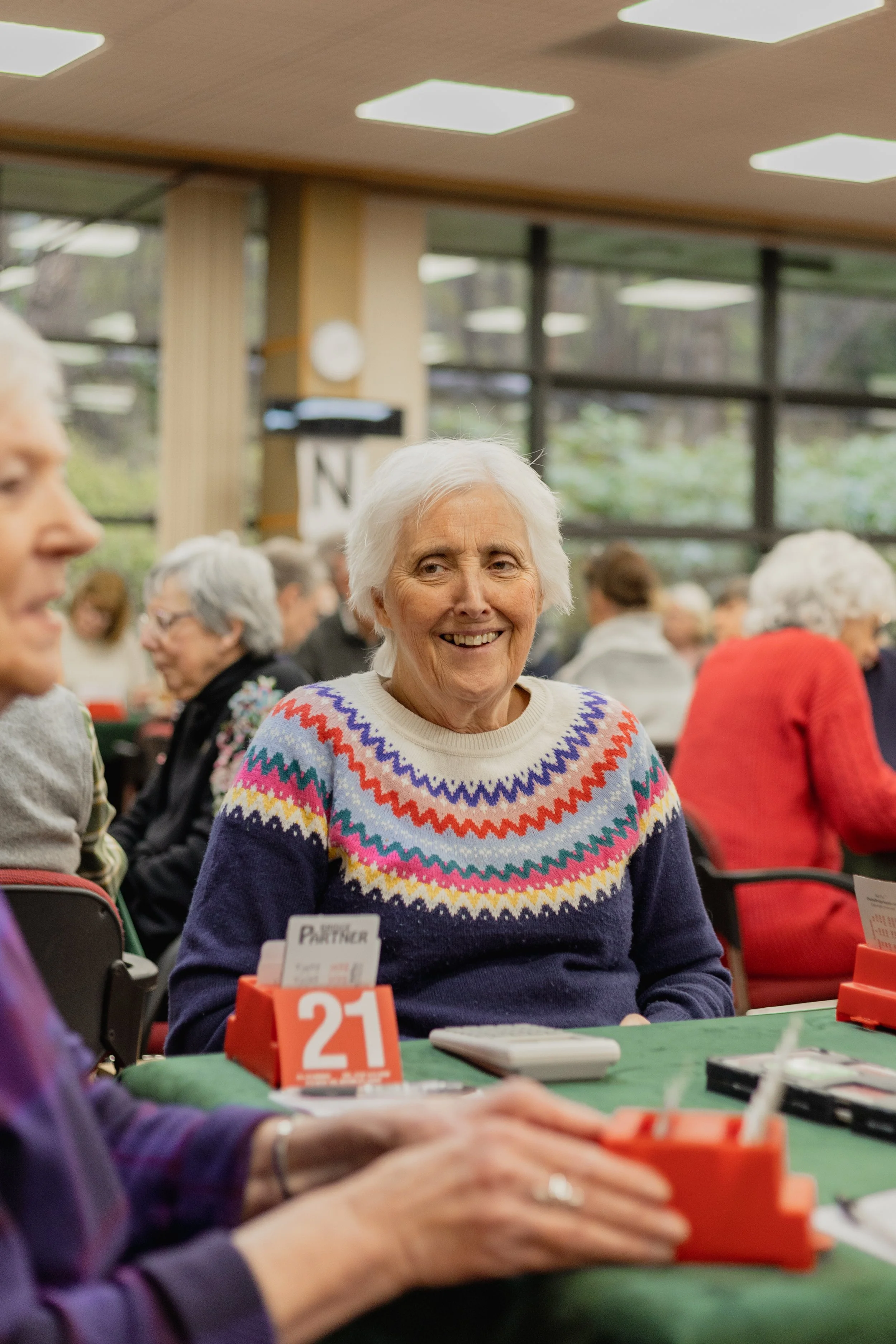 An elderly woman with white hair smiling while sitting at a table during a social gathering, wearing a multicolored patterned sweater.