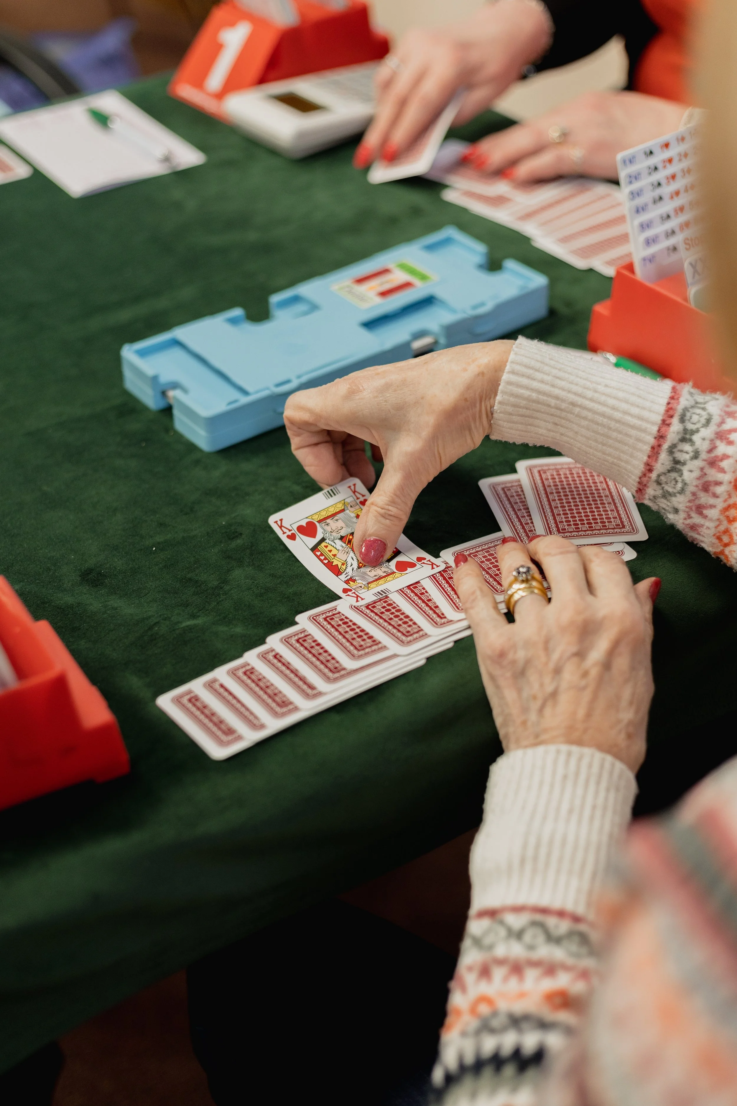 A close-up of an elderly woman's hands playing solitaire with red and white playing cards on a green table, with a blue card case and a score board nearby.