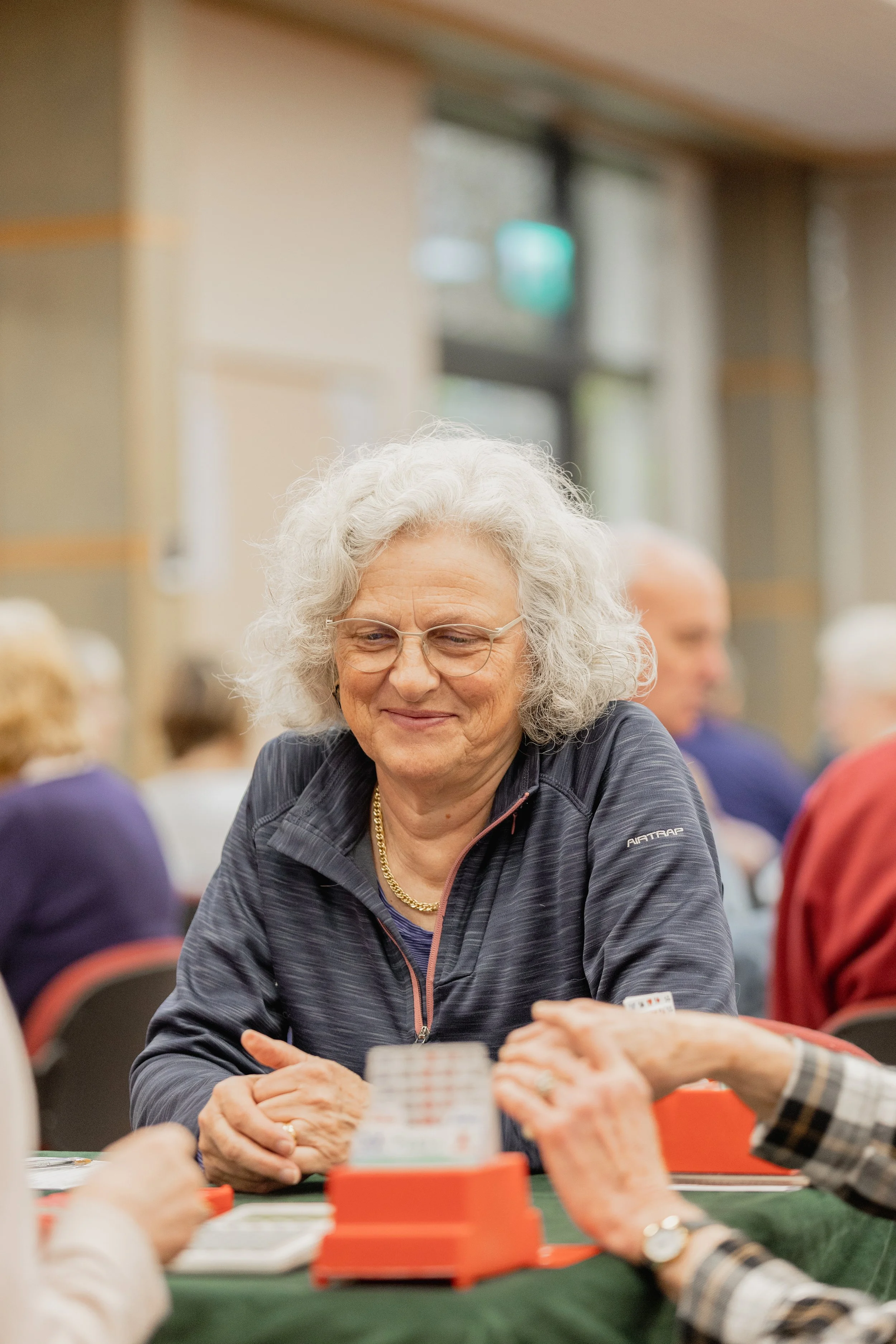 An elderly woman with curly gray hair and glasses playing a card game at a table with other people in a well-lit indoor setting.
