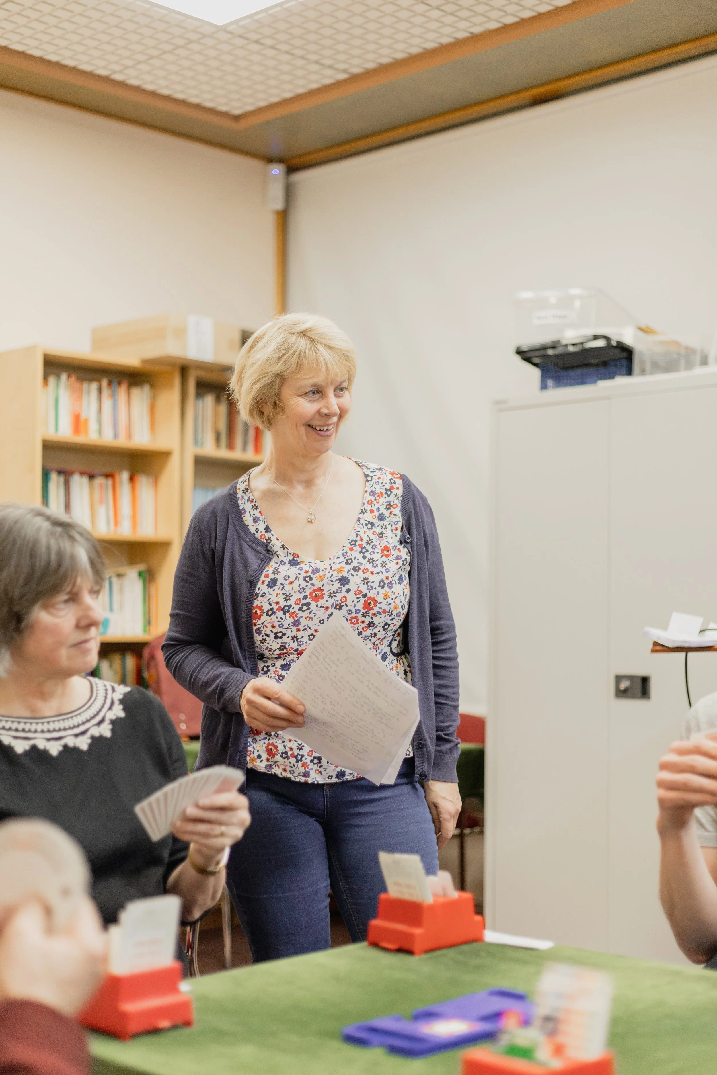 A woman with short blonde hair smiling, standing inside a room with bookshelves, holding a sheet of paper, surrounded by seated people holding papers or cards, with a setting that appears to be a community event or meeting.