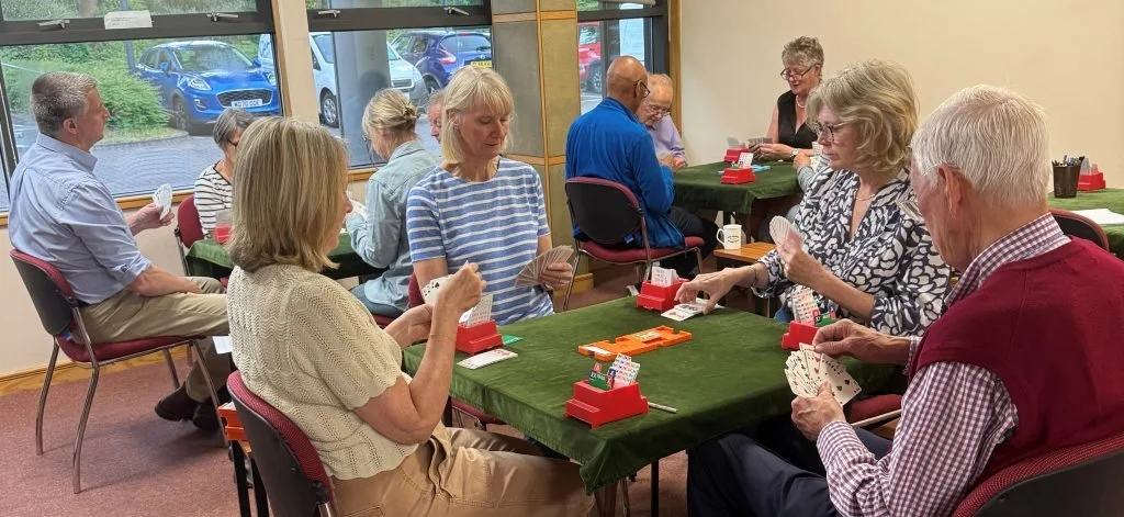 Group of older adults playing cards at tables in a room with large windows and parking lot view.