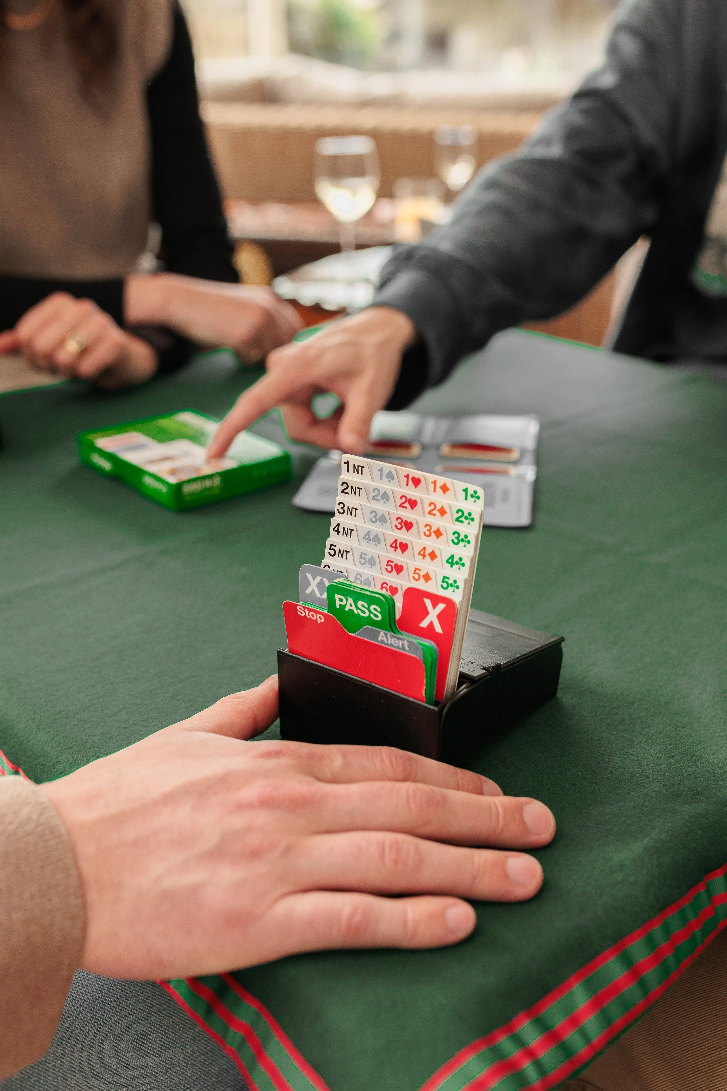 People playing a card game at a green-covered table with glasses of white wine in the background. A hand is handling a deck of action cards with symbols and descriptions like 'PASS' and 'Stop' placed in front of the player.