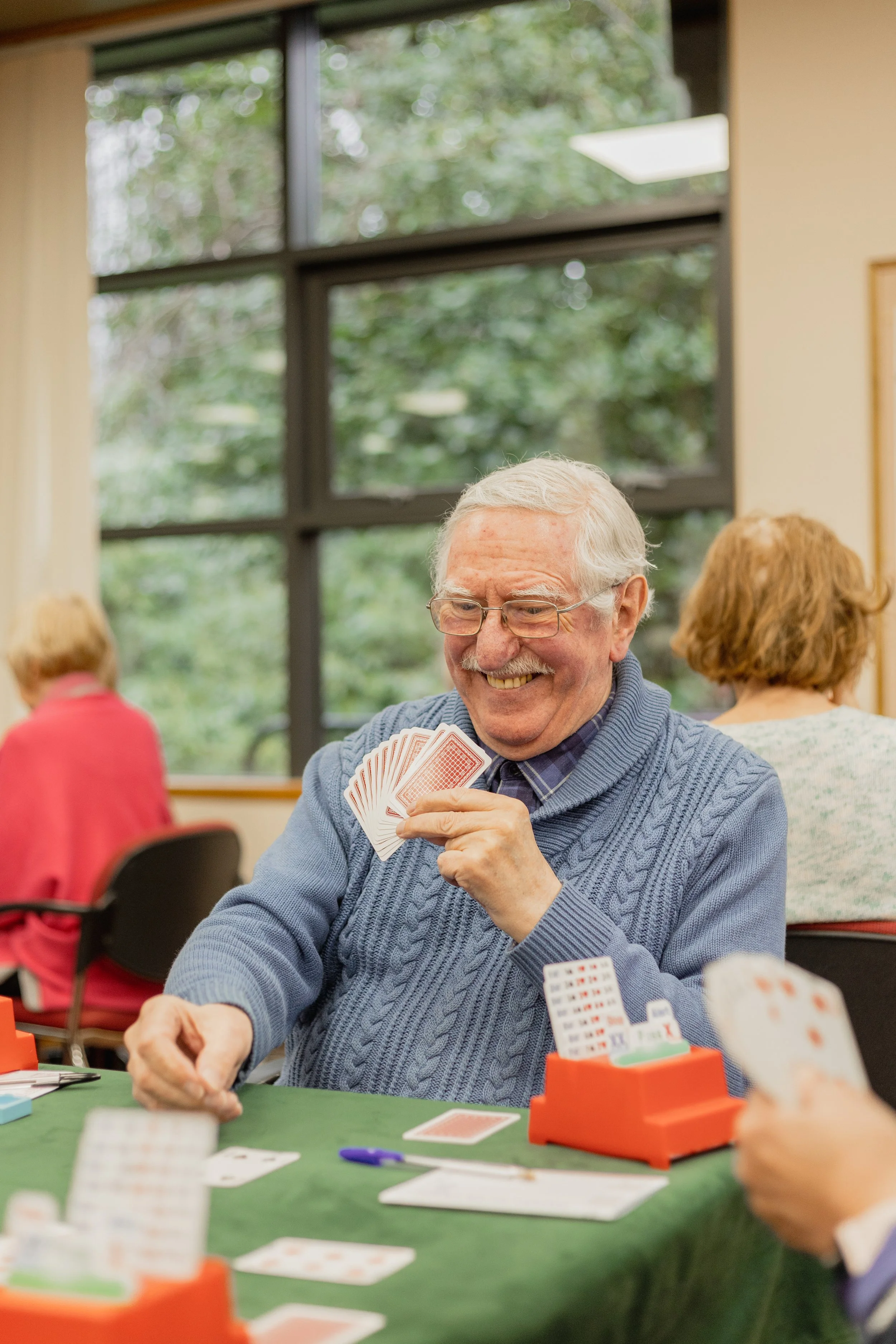An elderly man smiling and playing cards at a green felt table during a game with other people, with large windows and green trees outside in the background.