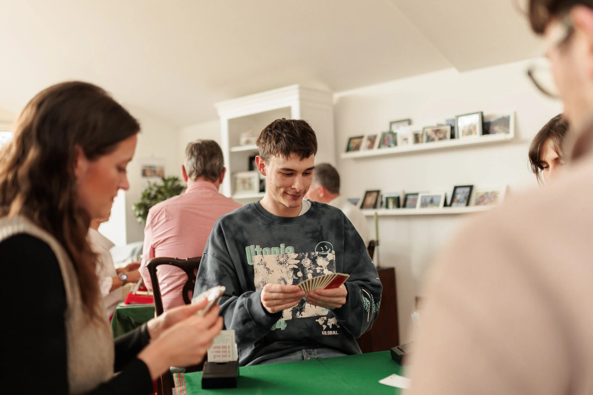 A young man sitting at a table playing a card game with a group of people in a living room, with framed photos on shelves in the background.