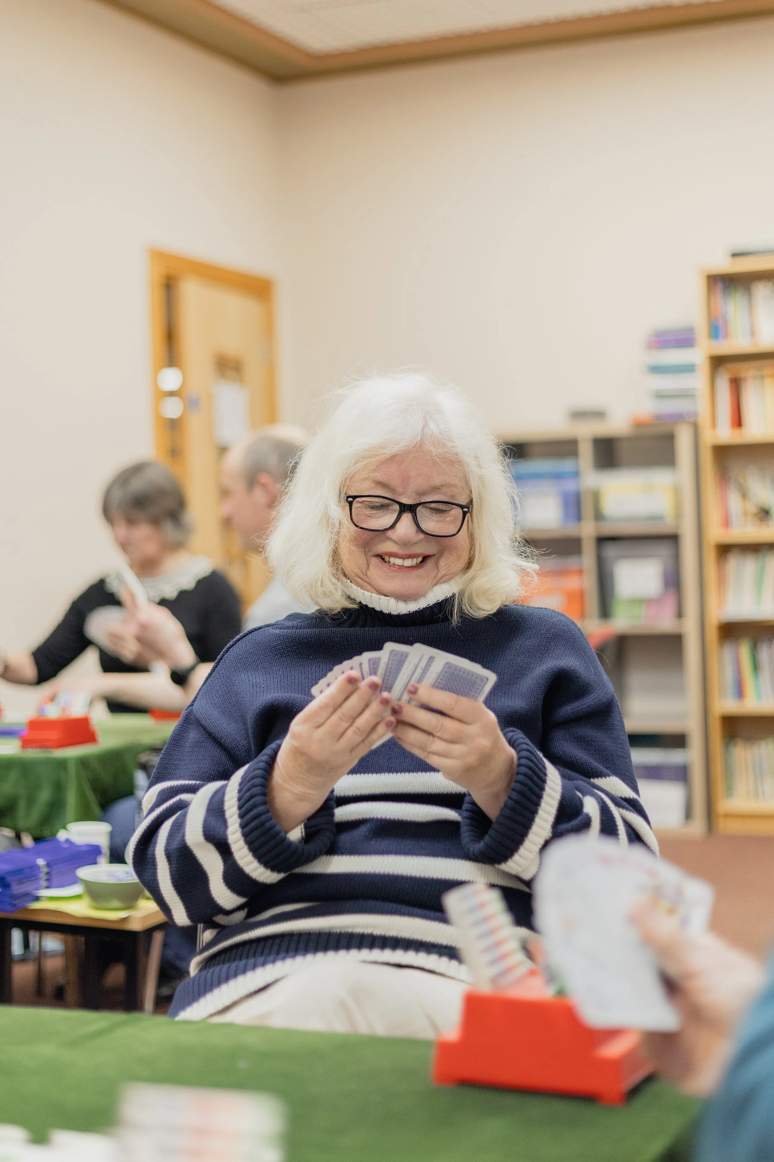 An elderly woman with glasses and white hair smiling and holding playing cards at a table in a room with bookshelves and other people in the background.