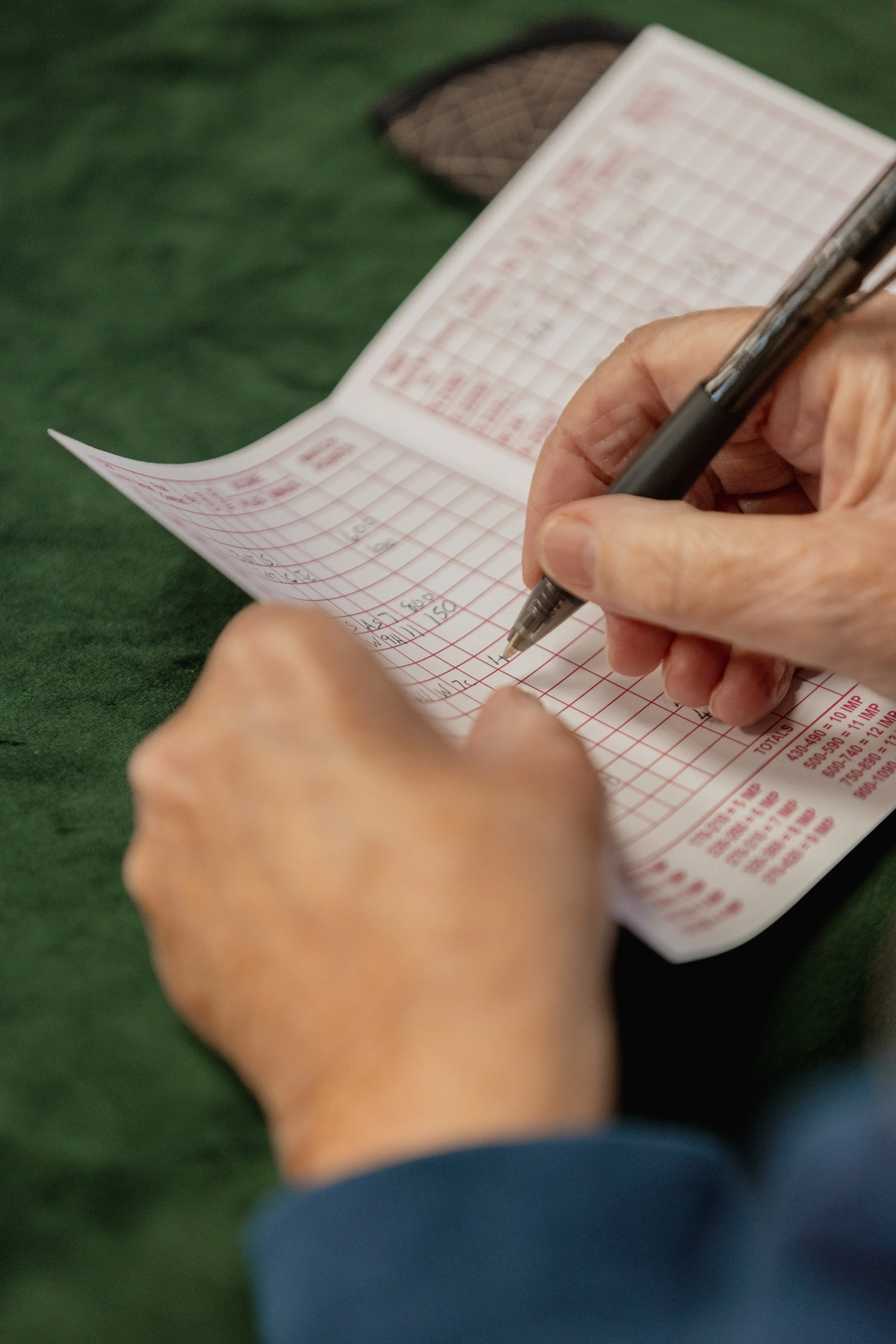 Close-up of a person's hands holding a pen and writing on a multi-column paper slip, likely a bingo card, on a dark green surface.