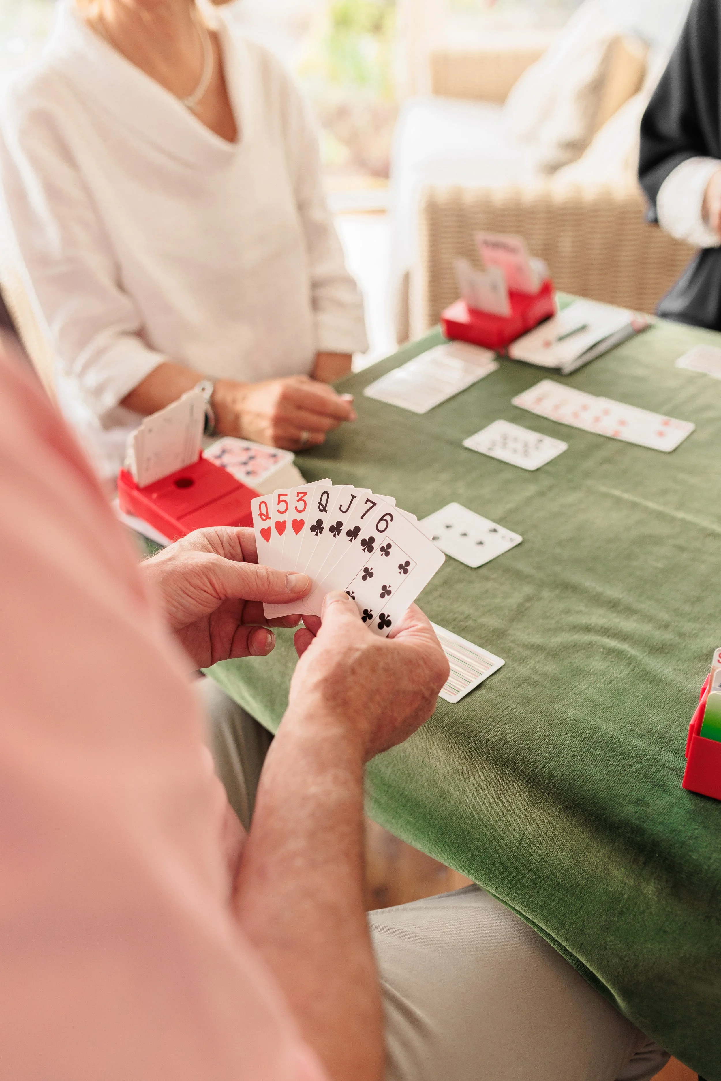 Person holding playing cards with Queen, 5, 3, Jack, 7, 6 of clubs, at a table with other playing cards and game pieces, in a well-lit room.