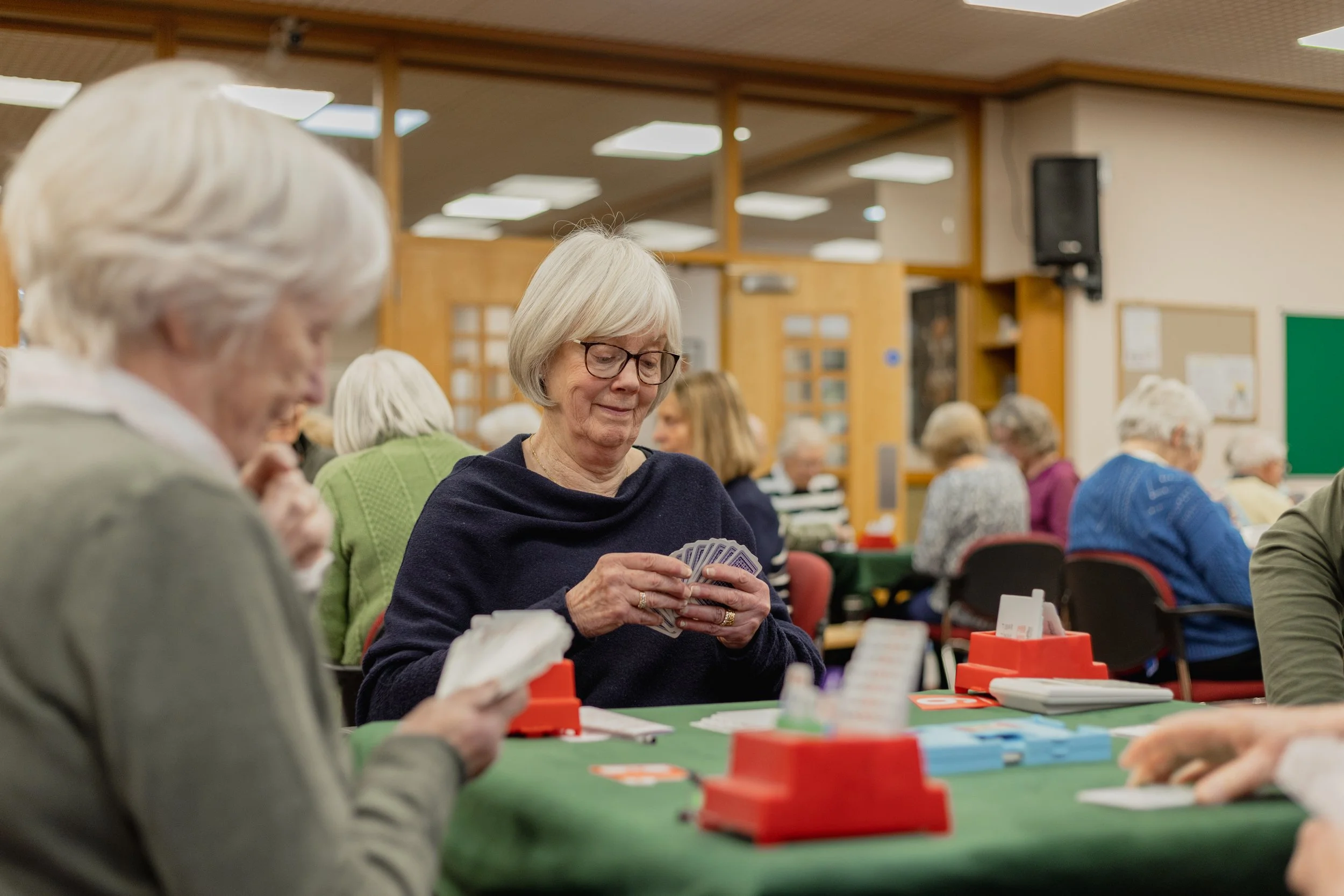 A group of elderly women playing cards at a social gathering in a community center with wooden paneling on the walls.