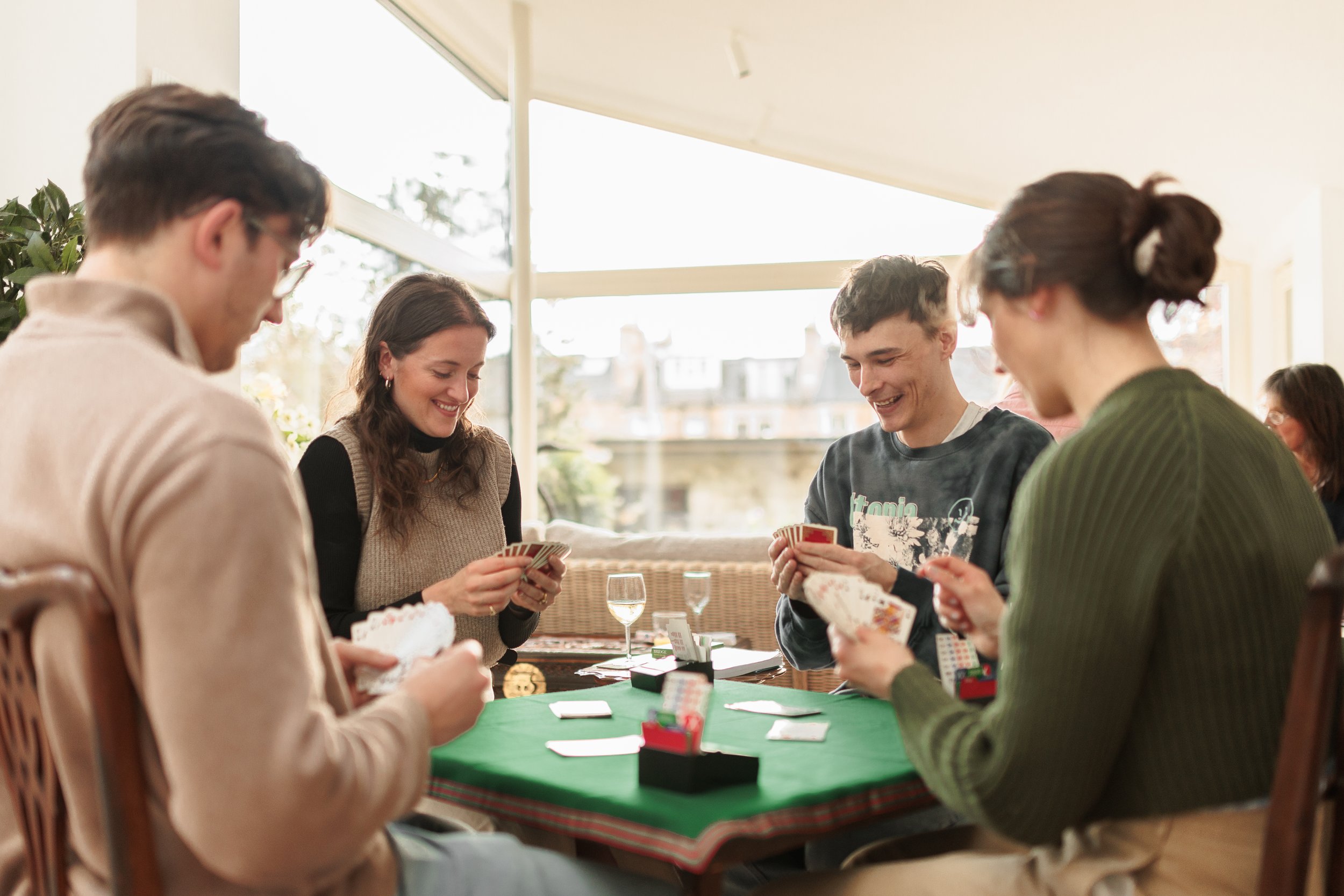 Group of five young adults playing cards around a table in a sunlit room.