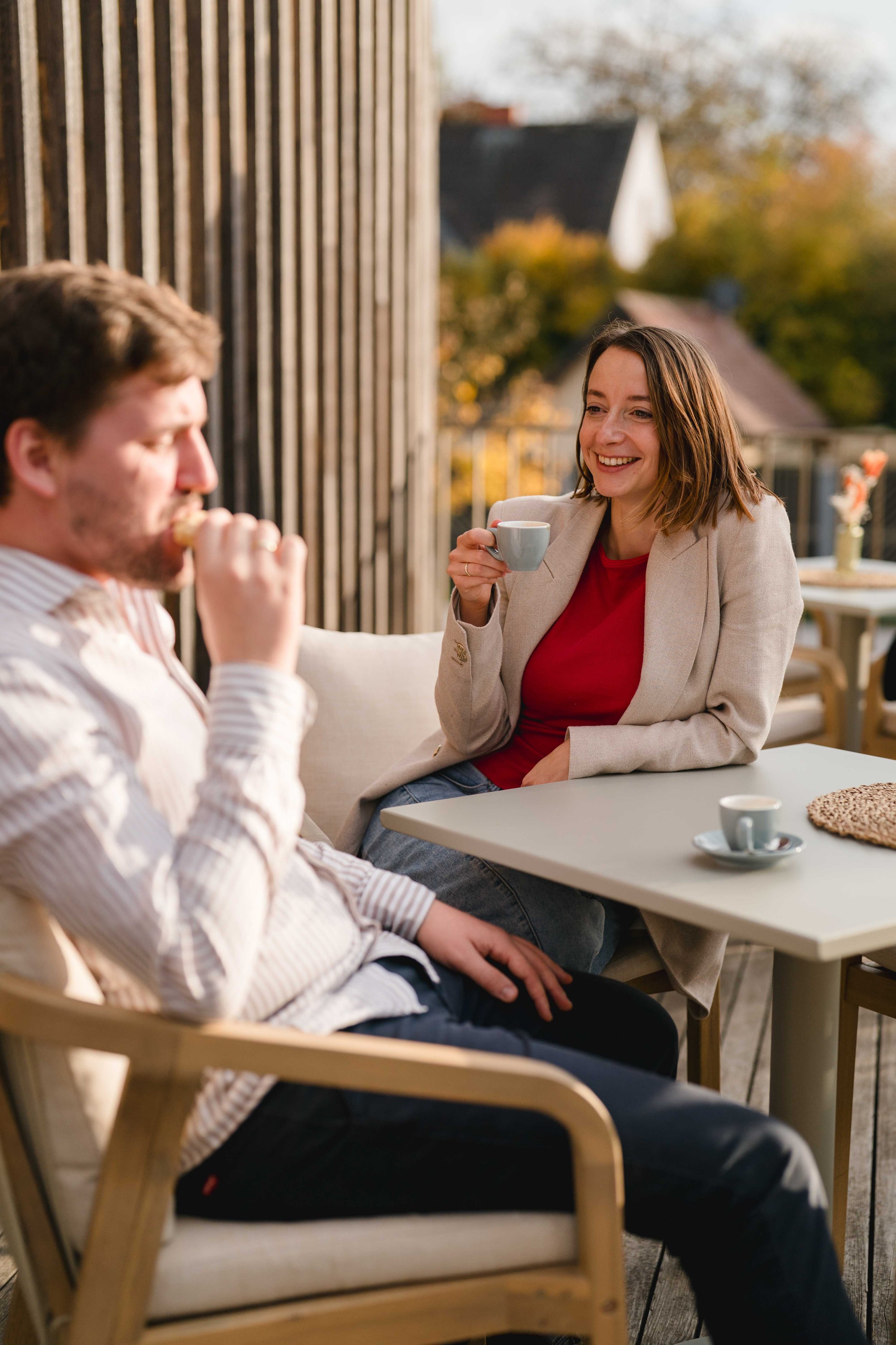 Zwei Menschen sitzen auf einer Terrasse, trinken Kaffee und unterhalten sich, umgeben von Herbstbäumen und Häusern im Hintergrund.