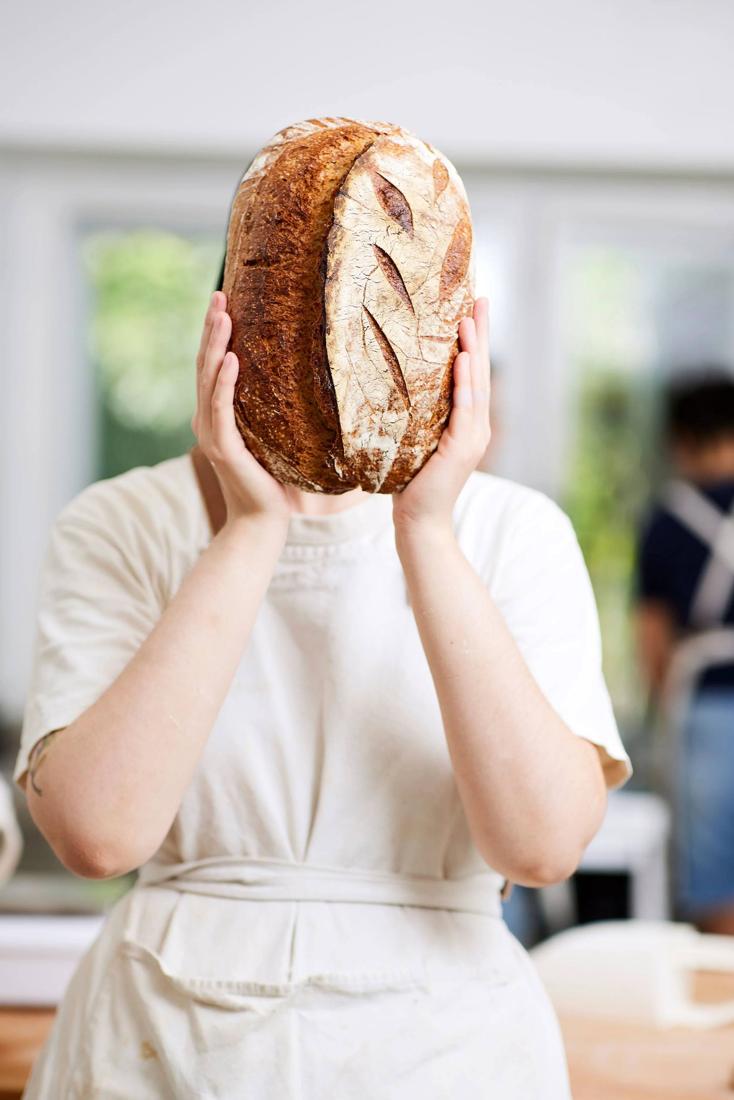 Frau hält ein großes, ovales, frisch gebackenes Brot vor ihr Gesicht, in einer hellen Küche.