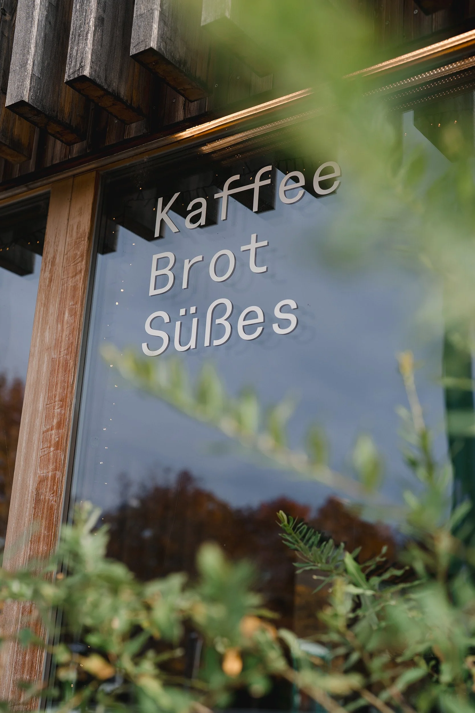 Die Scheibe eines Cafés mit der Aufschrift 'Kaffee Brot Süßes'; im Vordergrund befinden sich grüne Pflanzen, im Hintergrund ist Holzverkleidung und ein Fenster zu sehen.