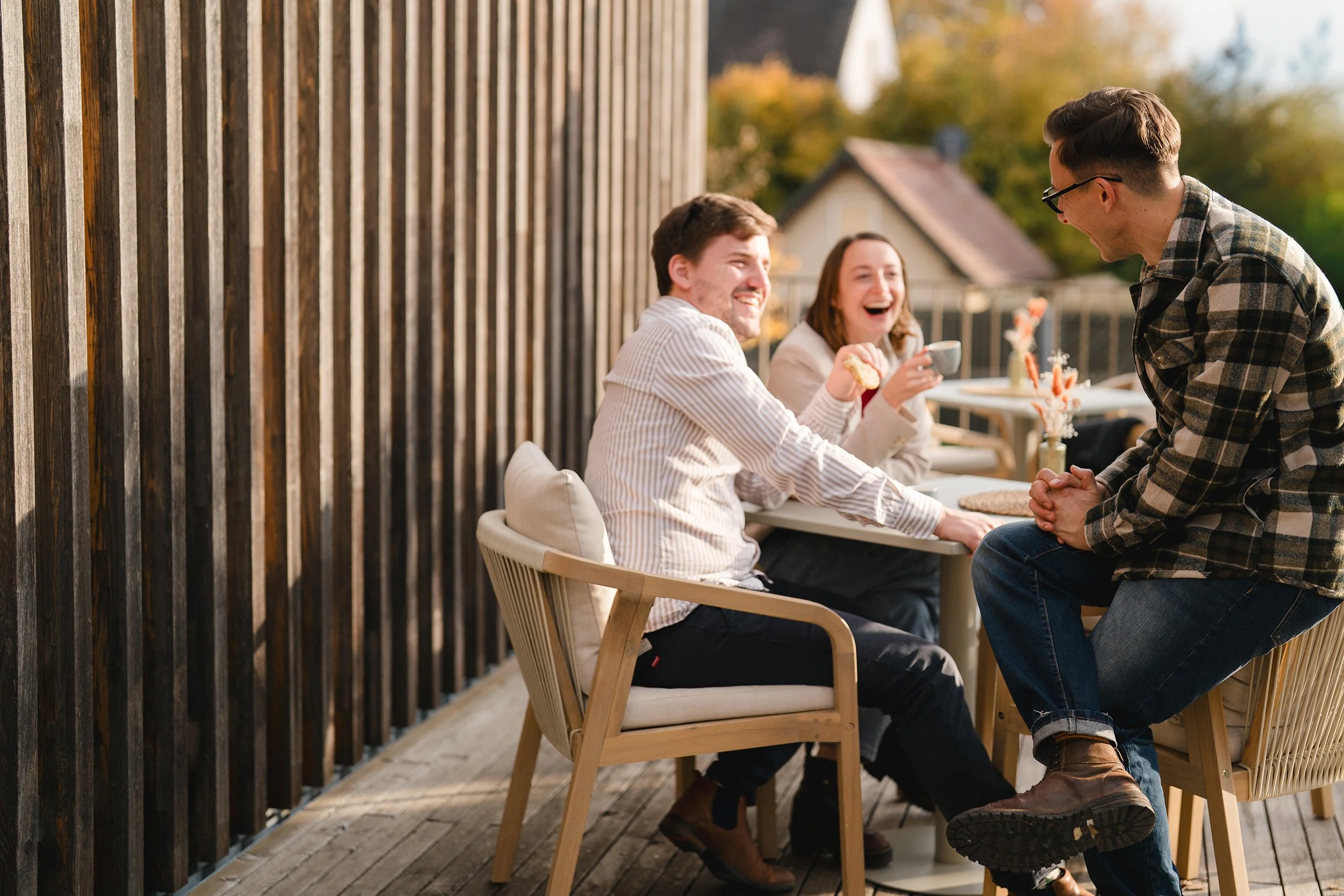 Vier junge Erwachsene sitzen und stehen auf einer Terrasse und lachen bei einem Treffen im Freien, während sie Kaffee trinken und sprechen.