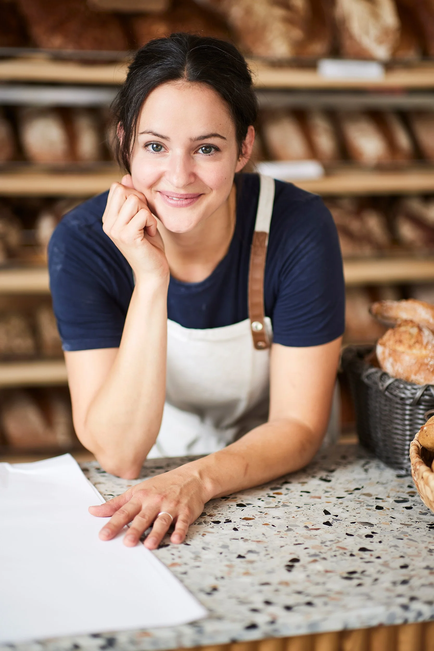 Eine junge Frau mit dunklem Haar, die in einer Bäckerei arbeitet, sitzt an einer Marmorarbeitsfläche mit Broten im Hintergrund.