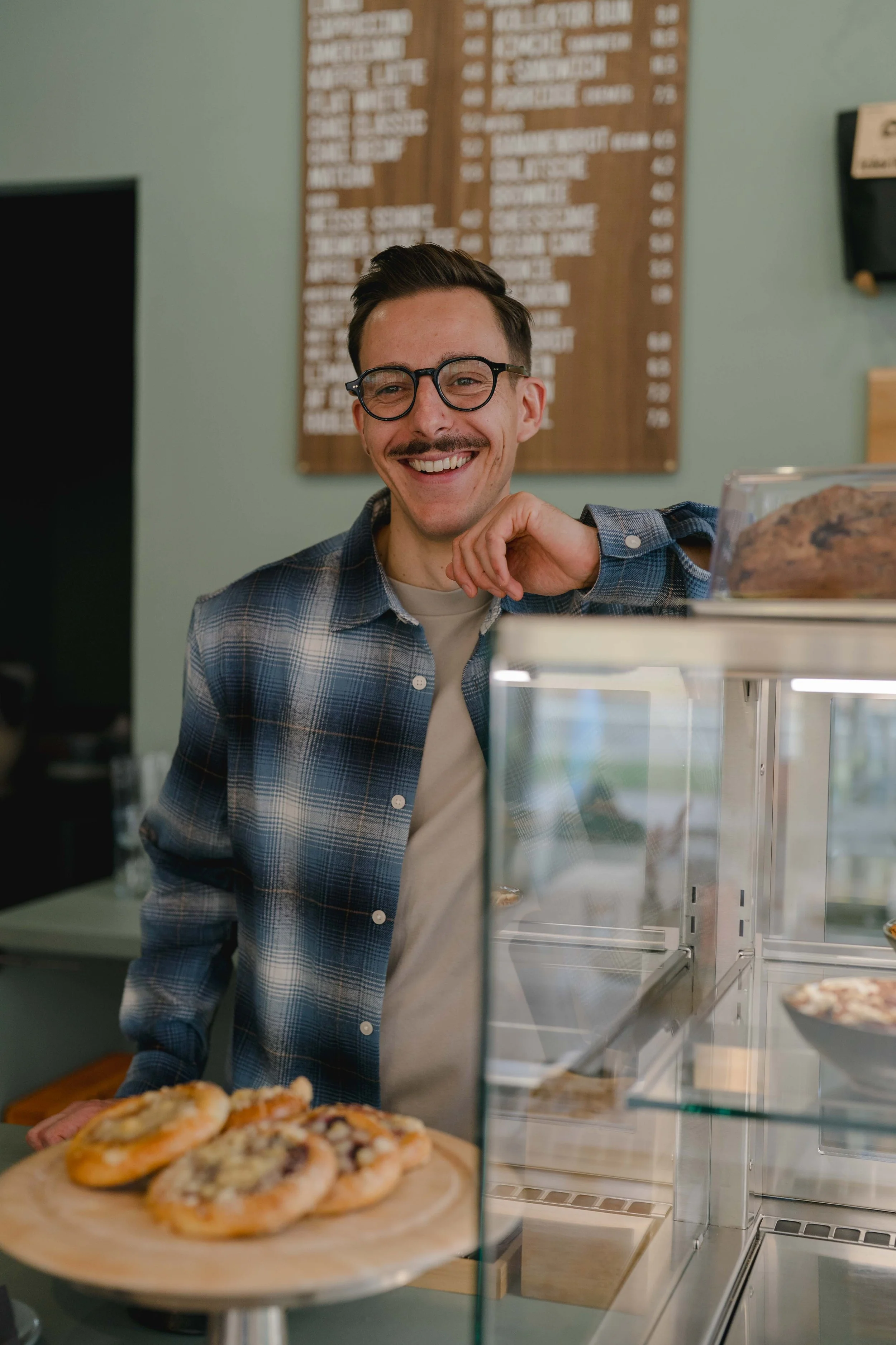 Ein junger Mann mit Brille und Schnurrbart steht hinter der Theke eines Pizzerias und lächelt, während Pizza auf einem Holzbrett präsentiert wird.