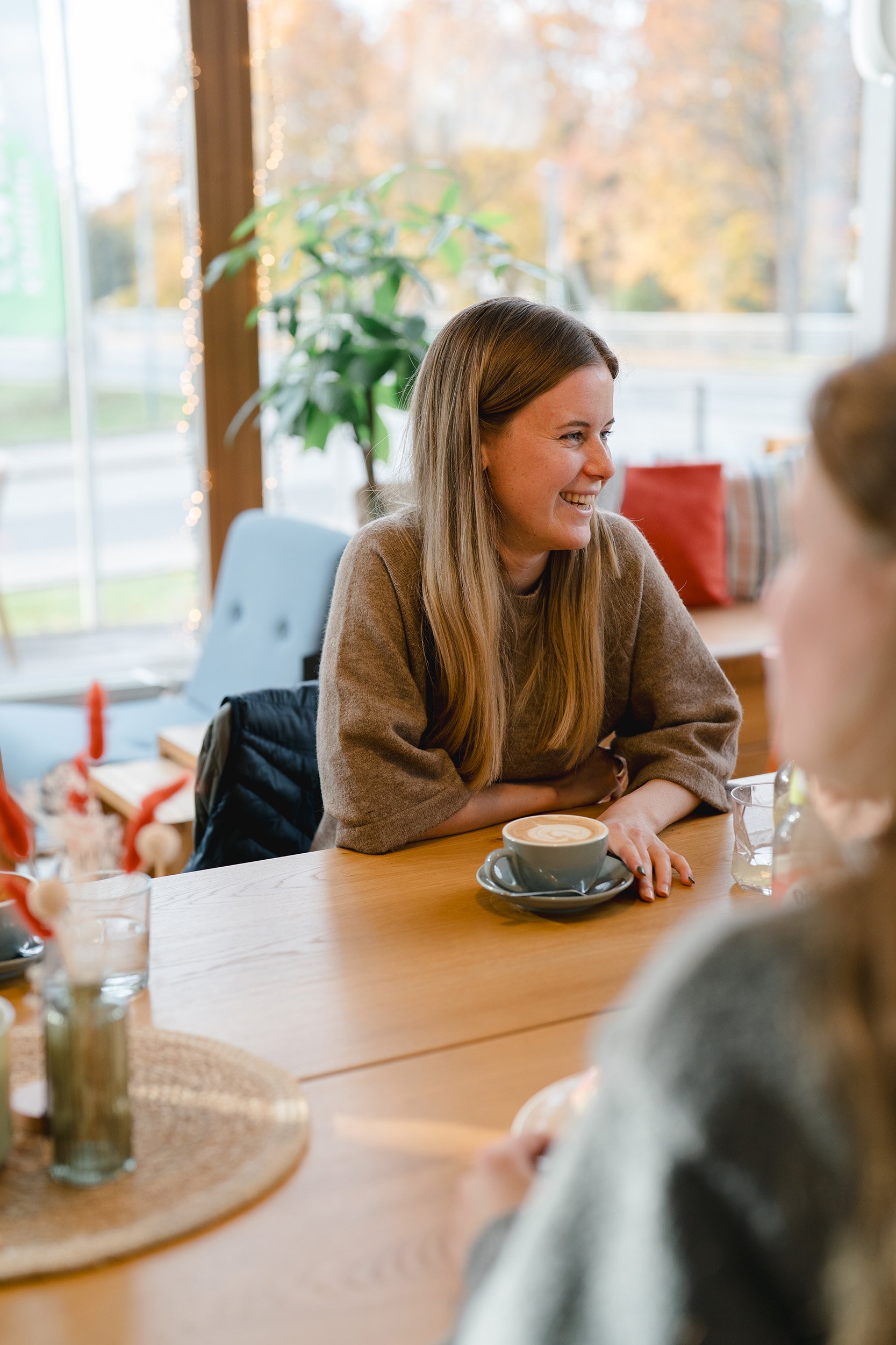 Eine lachende Frau mit langem braunem Haar sitzt an einem Tisch in einem gemütlichen Café, umgeben von Winterdeko, Cappuccino und Glas Wasser.