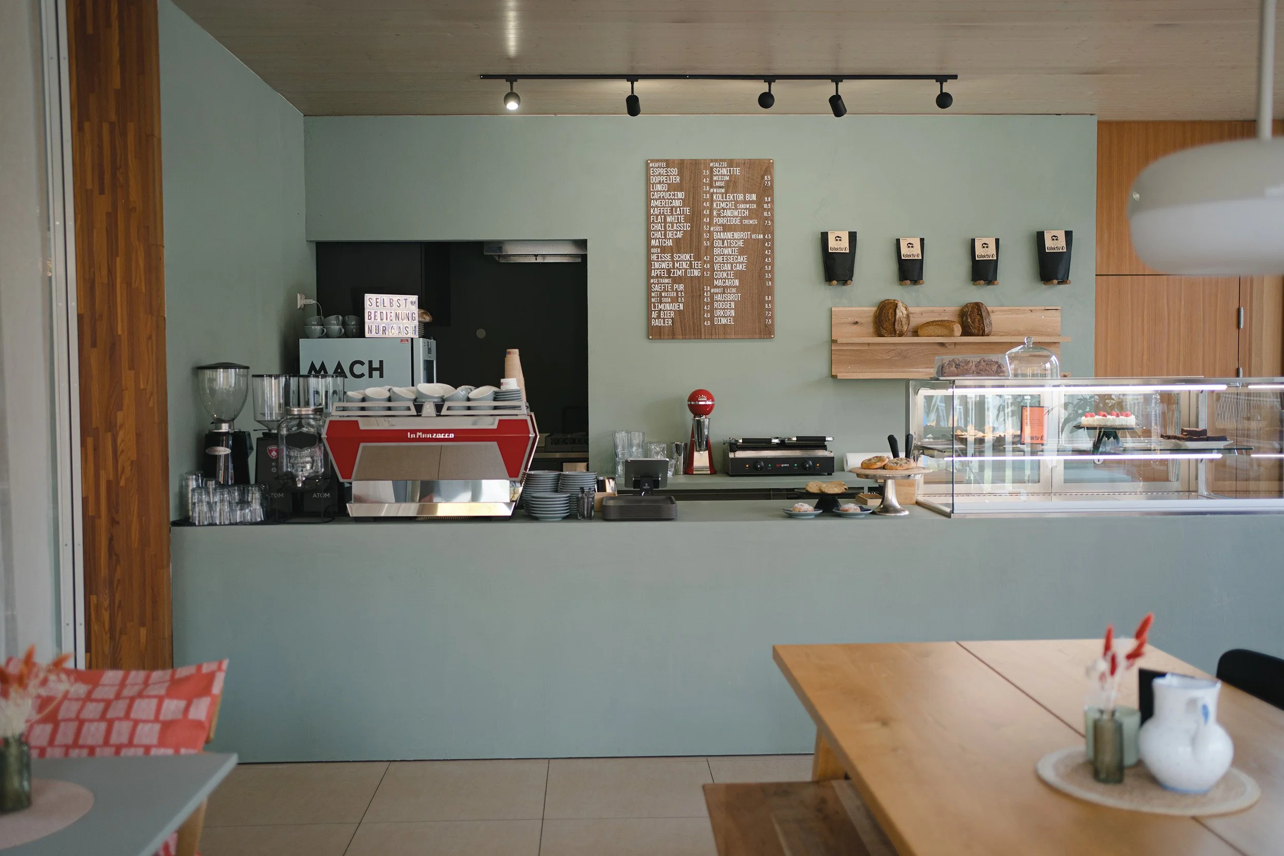 Inneneinrichtung eines Cafés mit Speisekarte an der Wand, Milch- und Kaffeemaschinen, Kuchen und Tassen auf der Theke, Holztisch im Vordergrund.