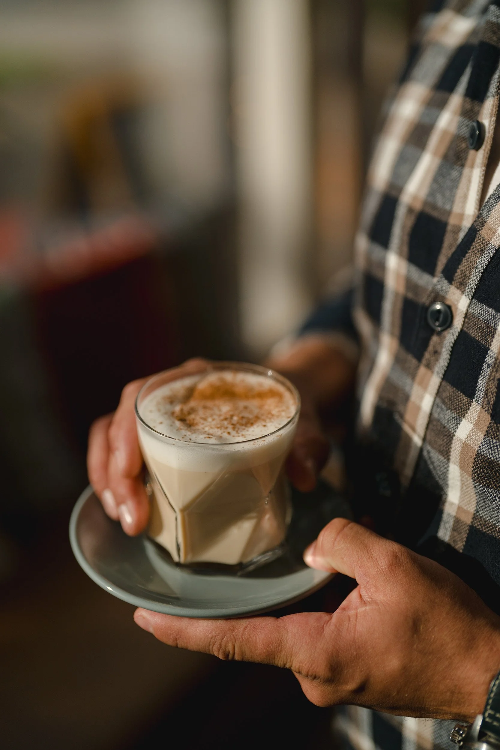 Eine Person in einem karierten Hemd hält ein Glas Cappuccino mit Milchschaum und Zimt auf einem kleinen weißen Teller.