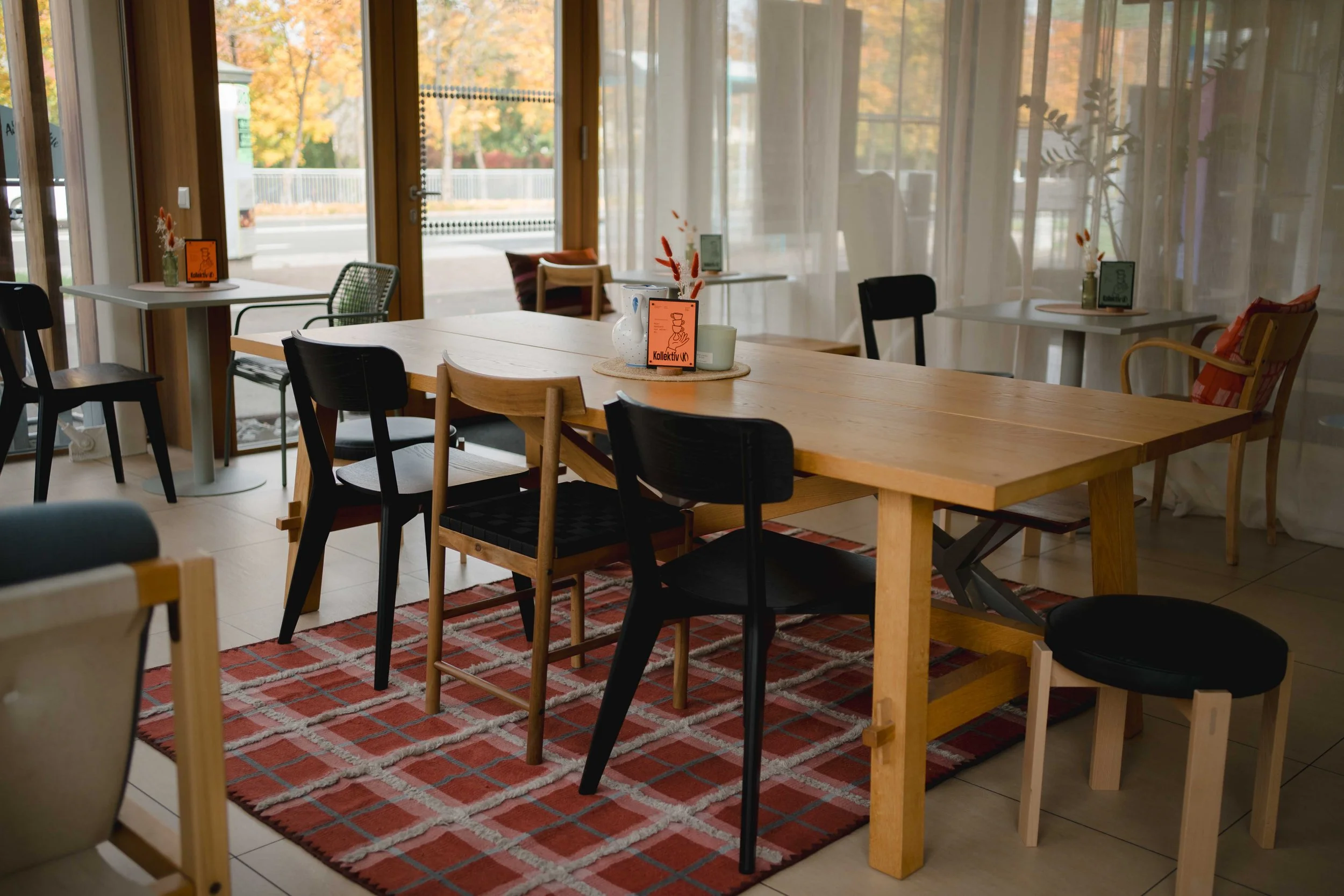 Innenraum eines gemütlichen Cafés mit mehreren Holztischen, bunten Stühlen und dekorativen Vasen mit Blumen. Große Fenster bieten Blick auf eine herbstliche Straße mit Bäumen in Orange und Gelb.