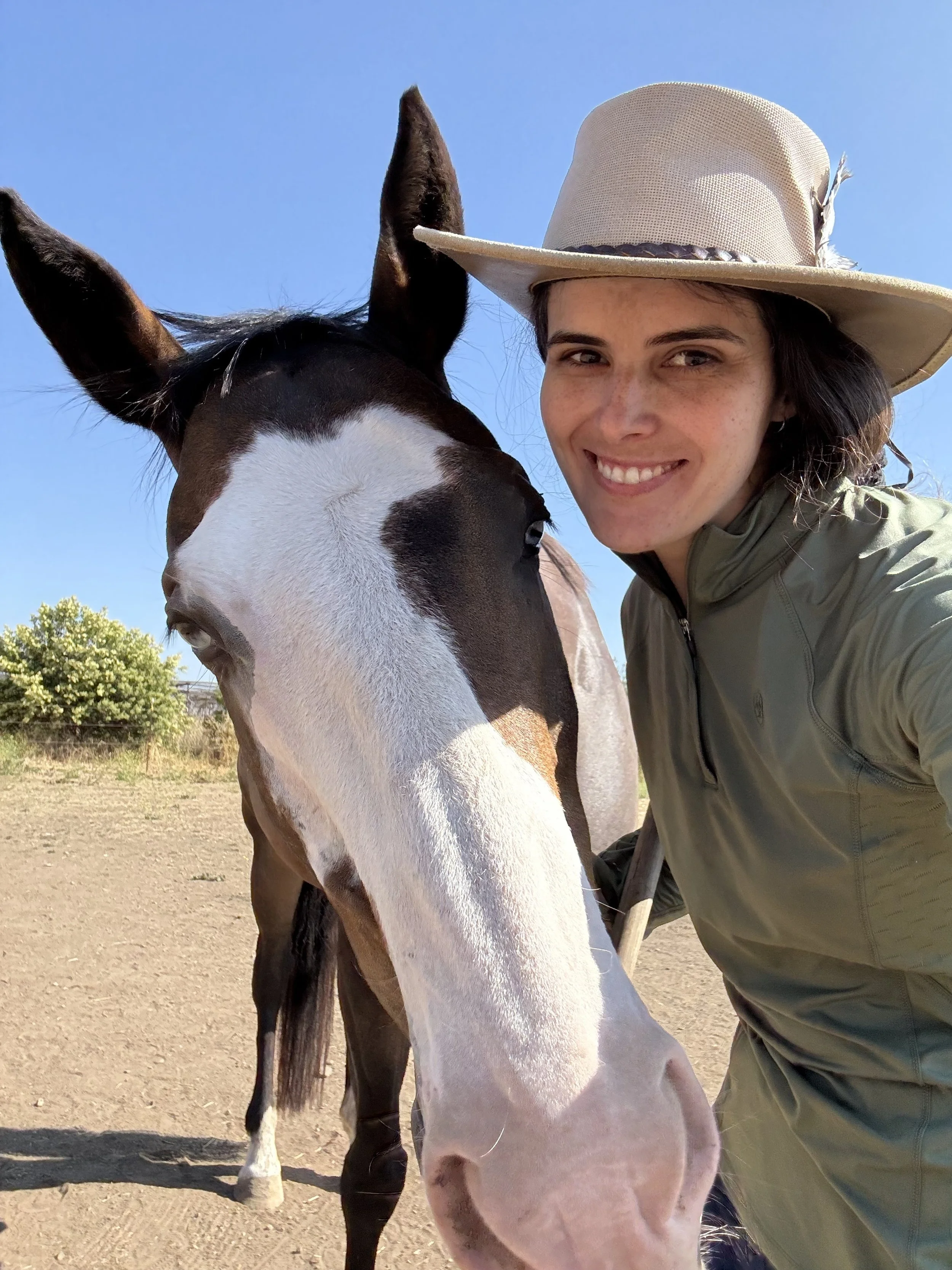 A woman with dark hair wearing a beige wide-brimmed hat and a green jacket taking a selfie with a black and white horse on a sunny day.