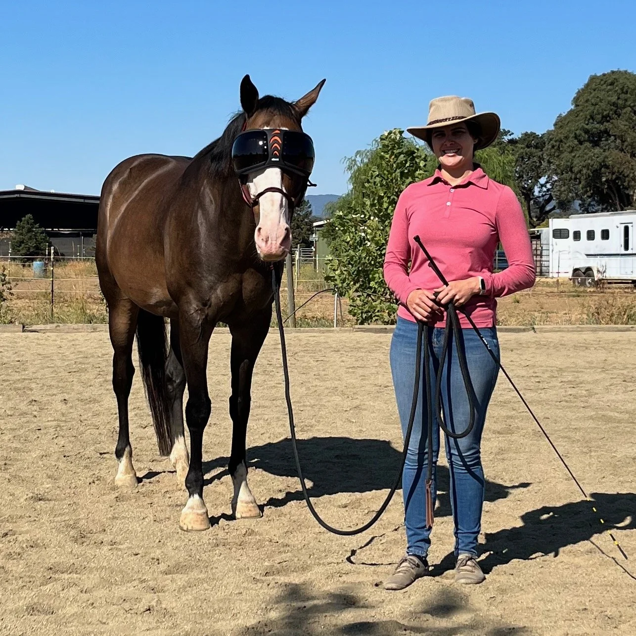 A woman standing next to a brown horse wearing a black riding mask, holding a lead rope. The woman is wearing a pink long-sleeve shirt, blue jeans, gray sneakers, and a beige wide-brim hat, smiling in an outdoor enclosed riding arena.